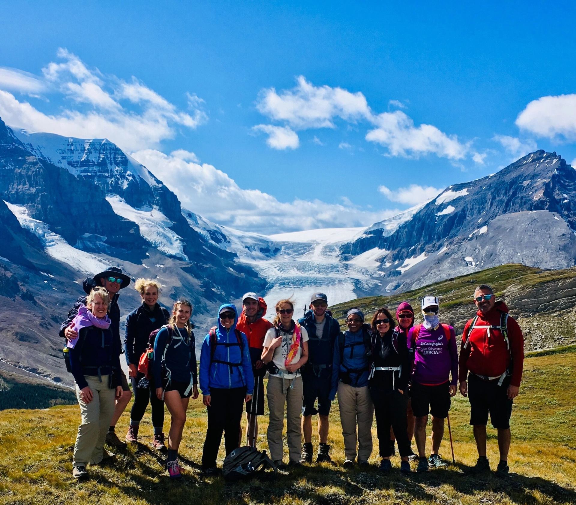 Group of hikers posing on grassy ridge with glacier and snow-capped mountains behind.