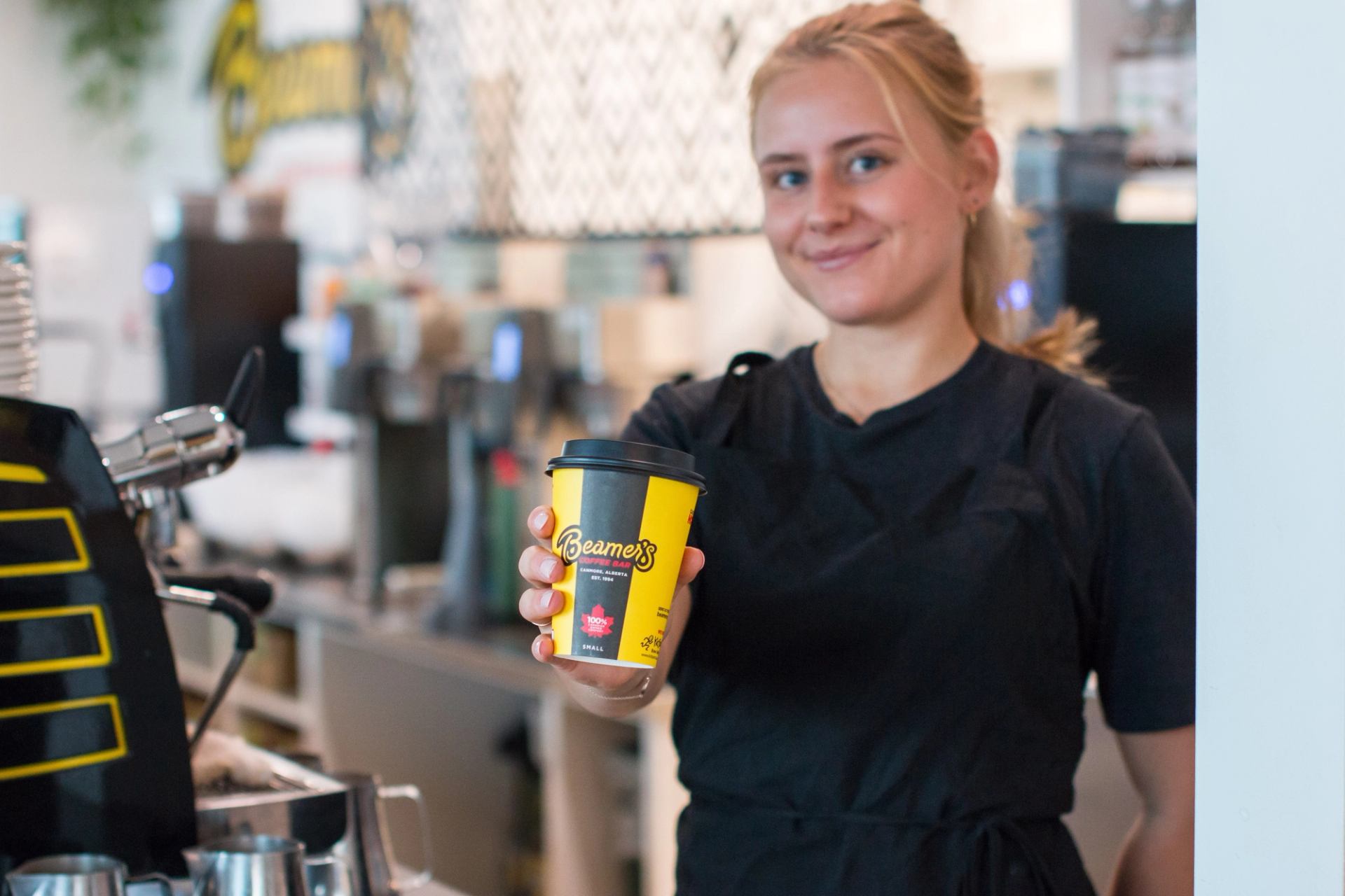 Person holding a yellow Beamer’s Coffee Bar cup behind the counter near an espresso machine.