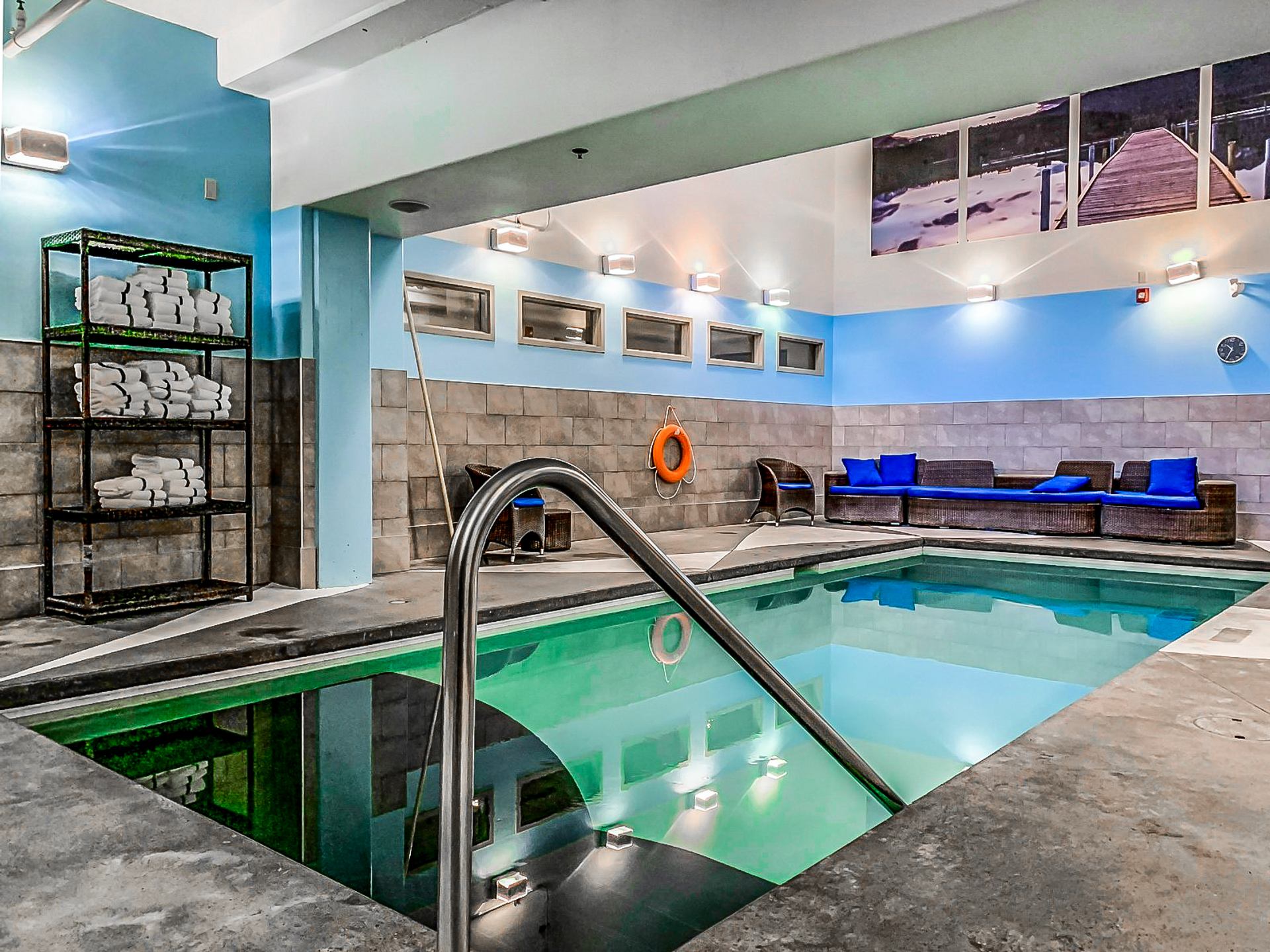 Indoor swimming pool with metal handrail, towel shelves, wicker seating and blue cushions.