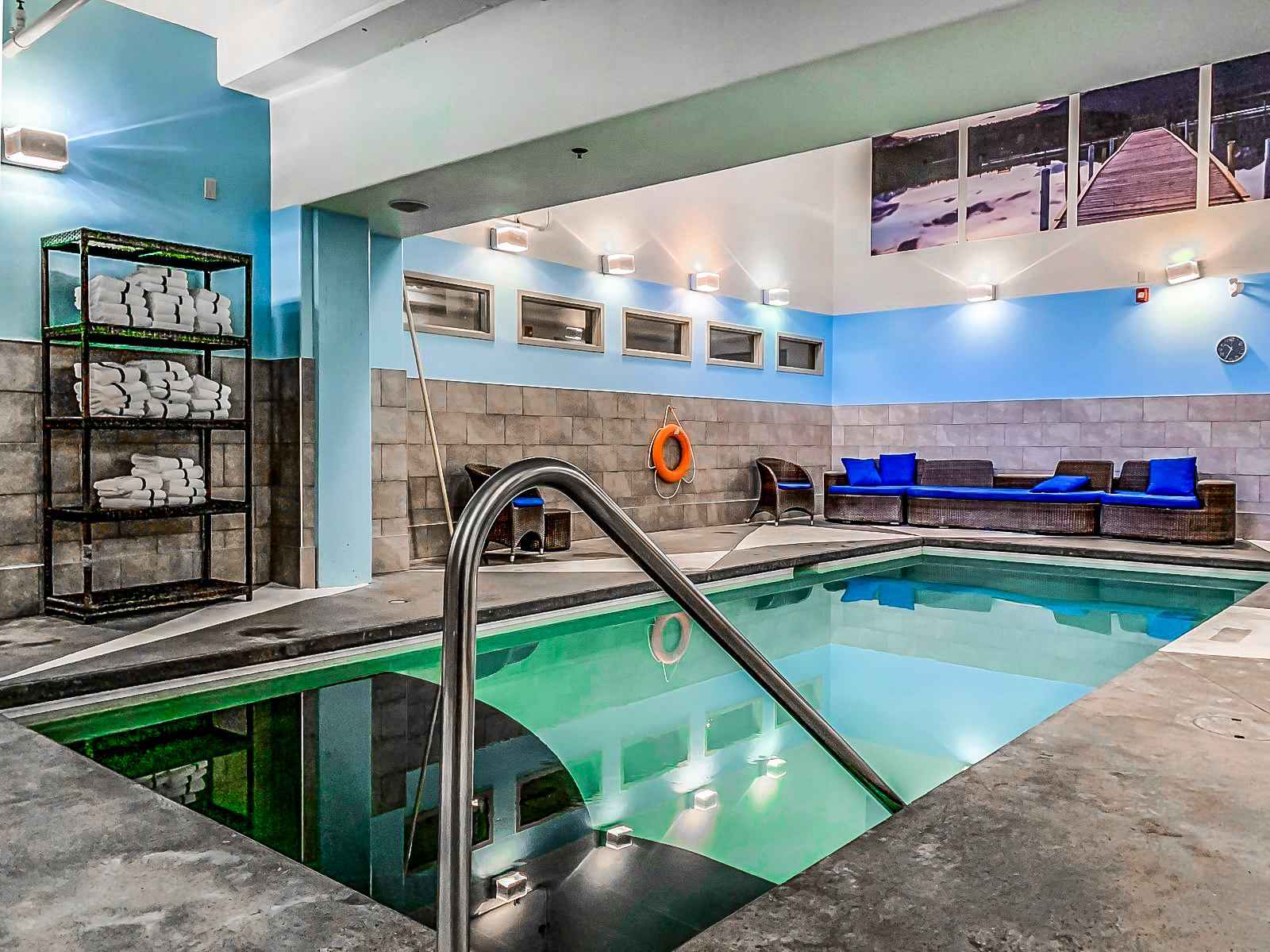 Indoor swimming pool with metal handrail, towel shelves, wicker seating and blue cushions.