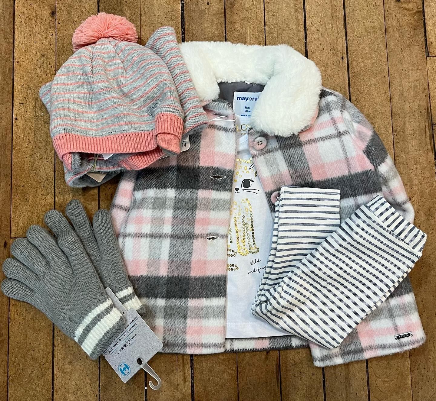 Kids winter clothing display: striped shirts, pink hat, grey gloves on wooden wall.