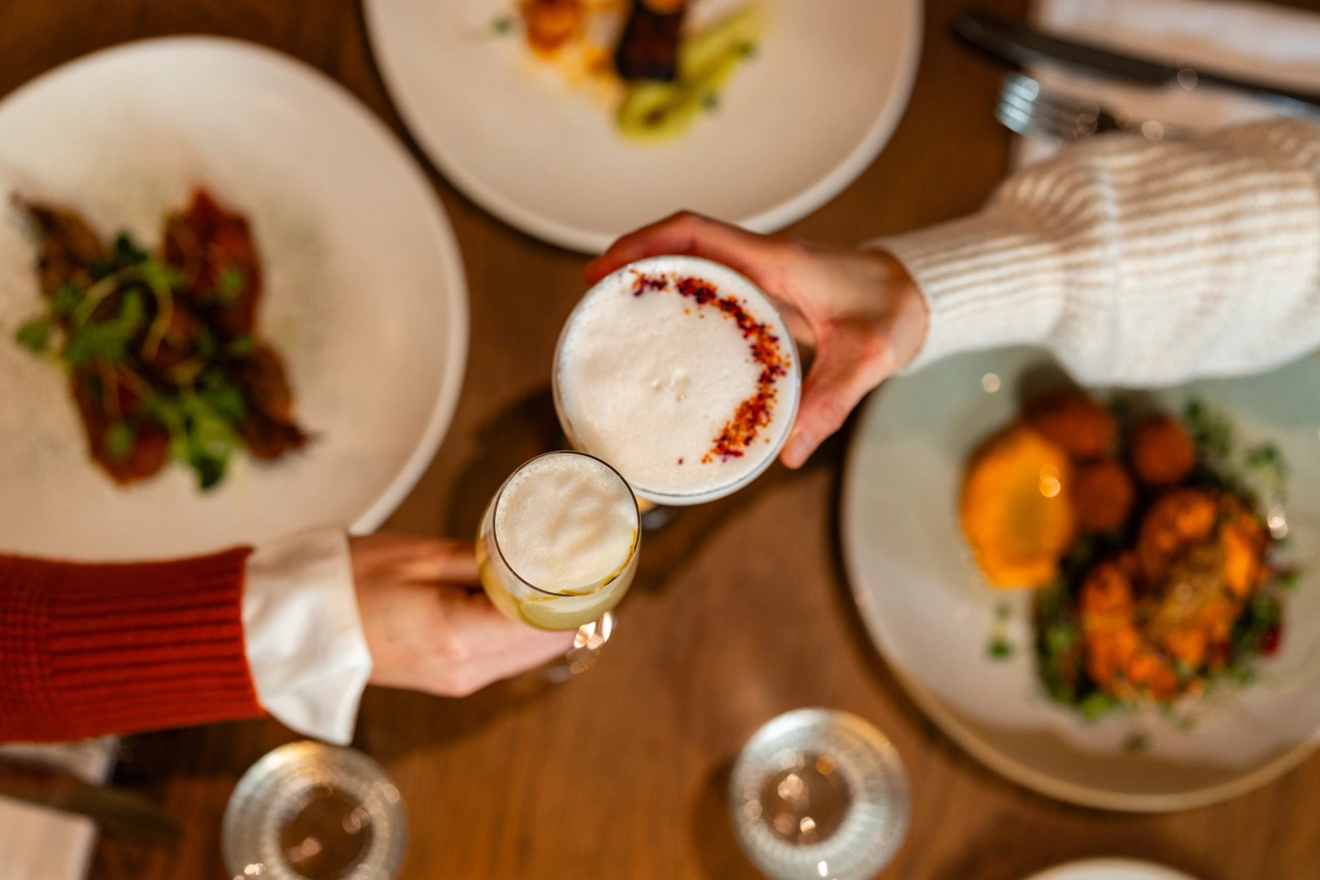 Two diners holding artisan cocktails over a table of food at Good Folk Banff