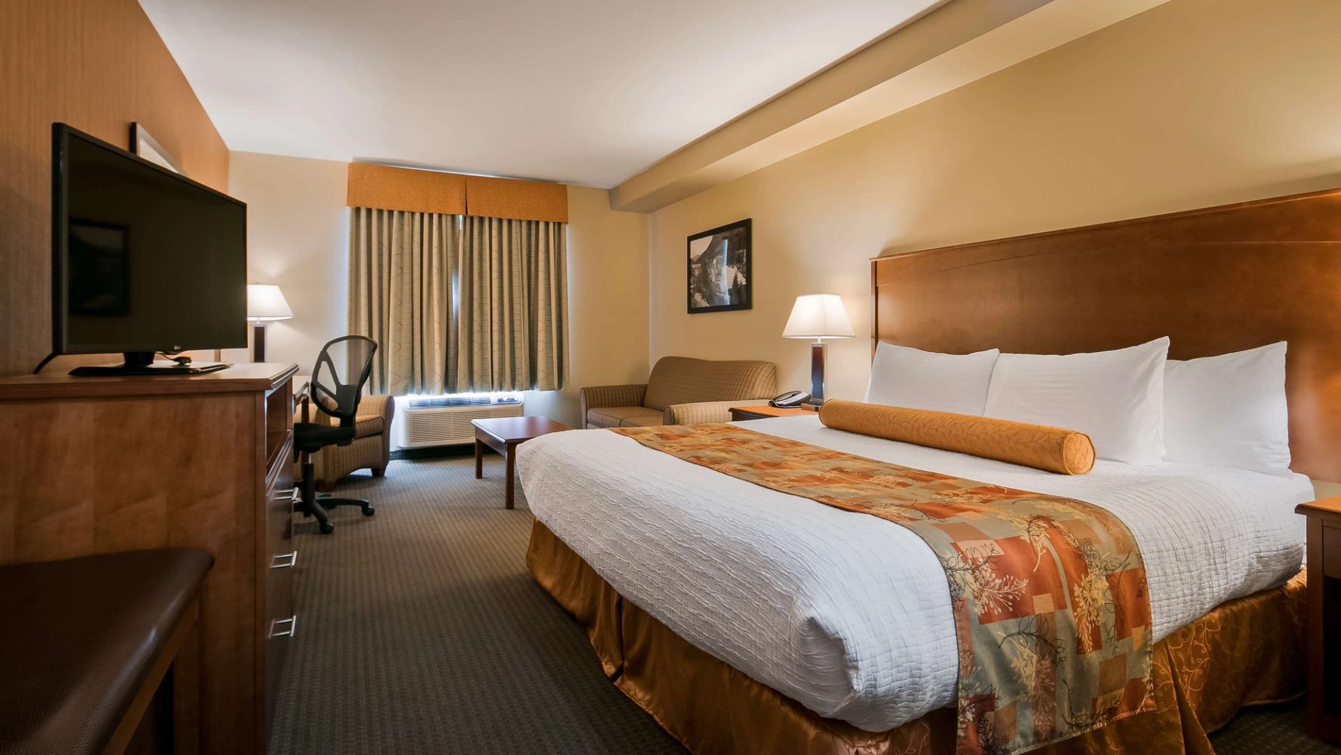 Hotel room with king bed, TV, desk chair, sofa, lamps and curtained window.