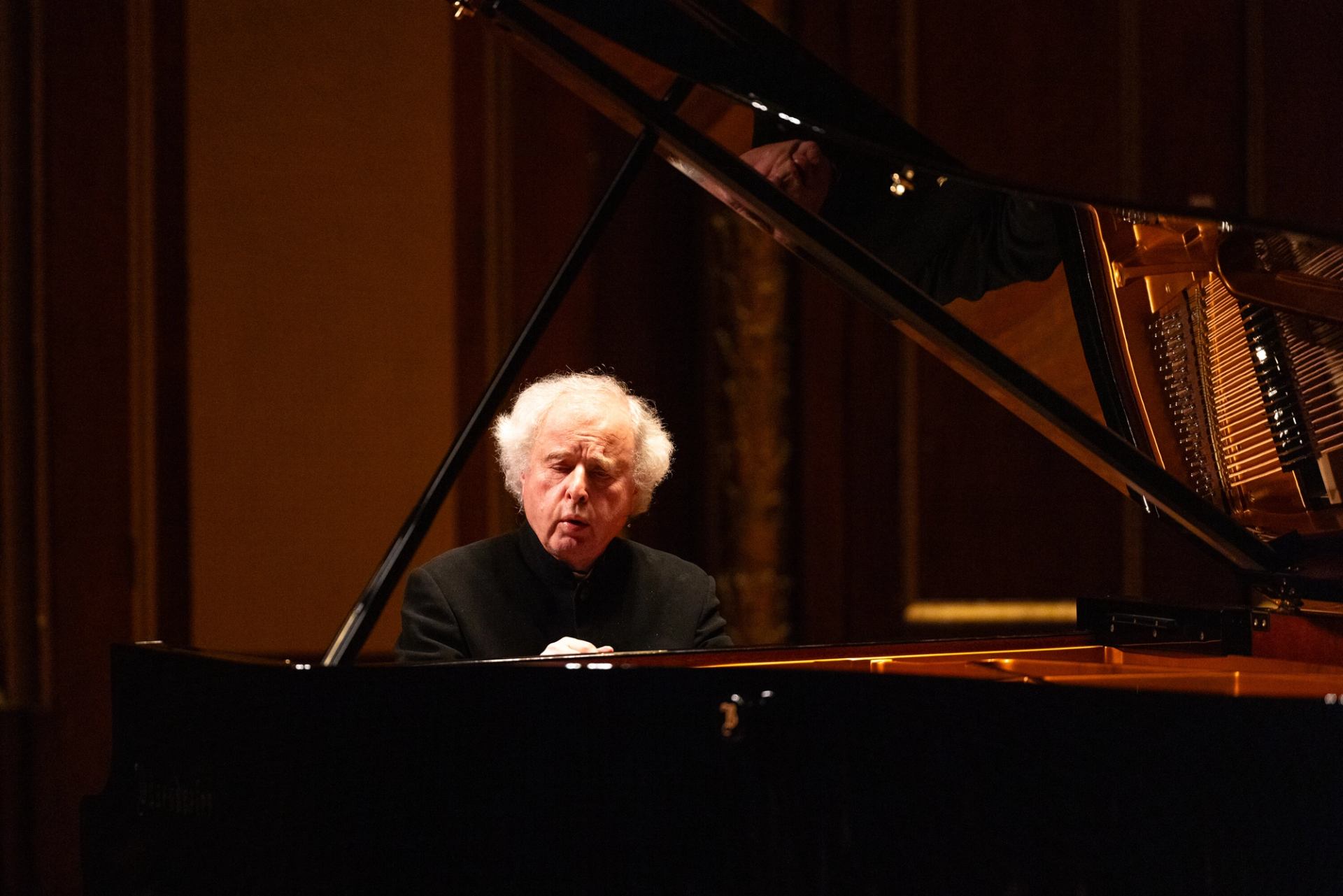 Pianist with white hair plays grand piano on dimly lit concert hall stage.