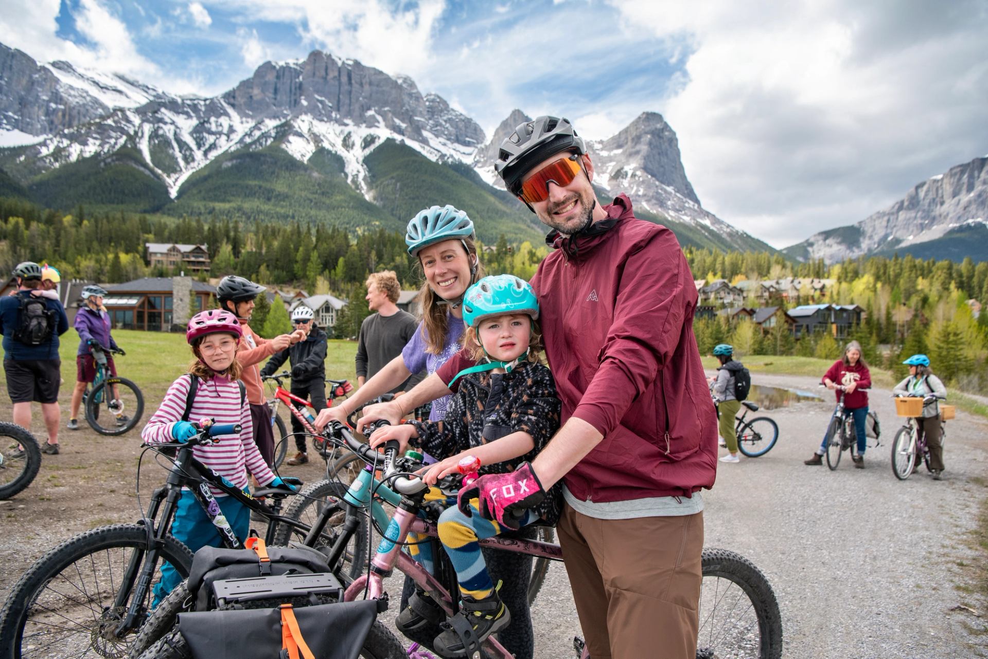 Cyclists with children on bikes gather on gravel path below snow-capped mountain range.