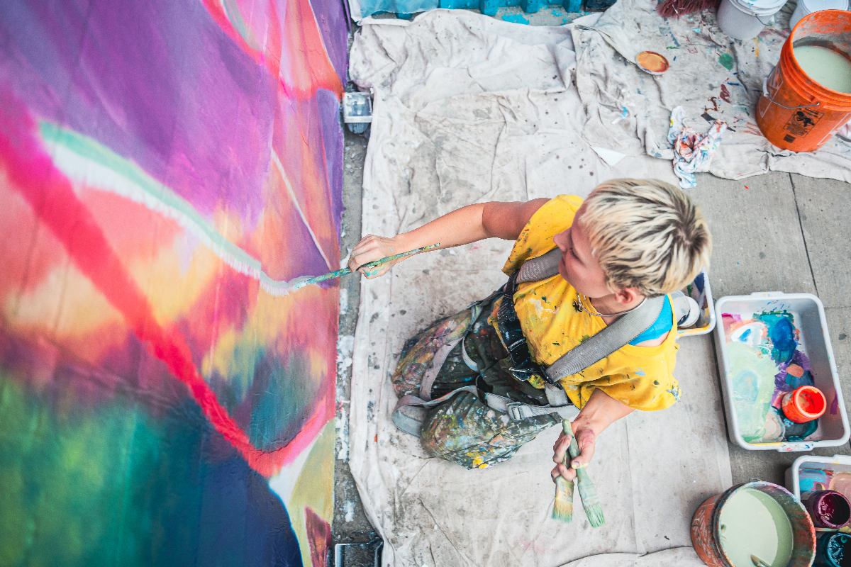 Person in yellow shirt paints a vibrant abstract mural, surrounded by paint supplies on the floor.