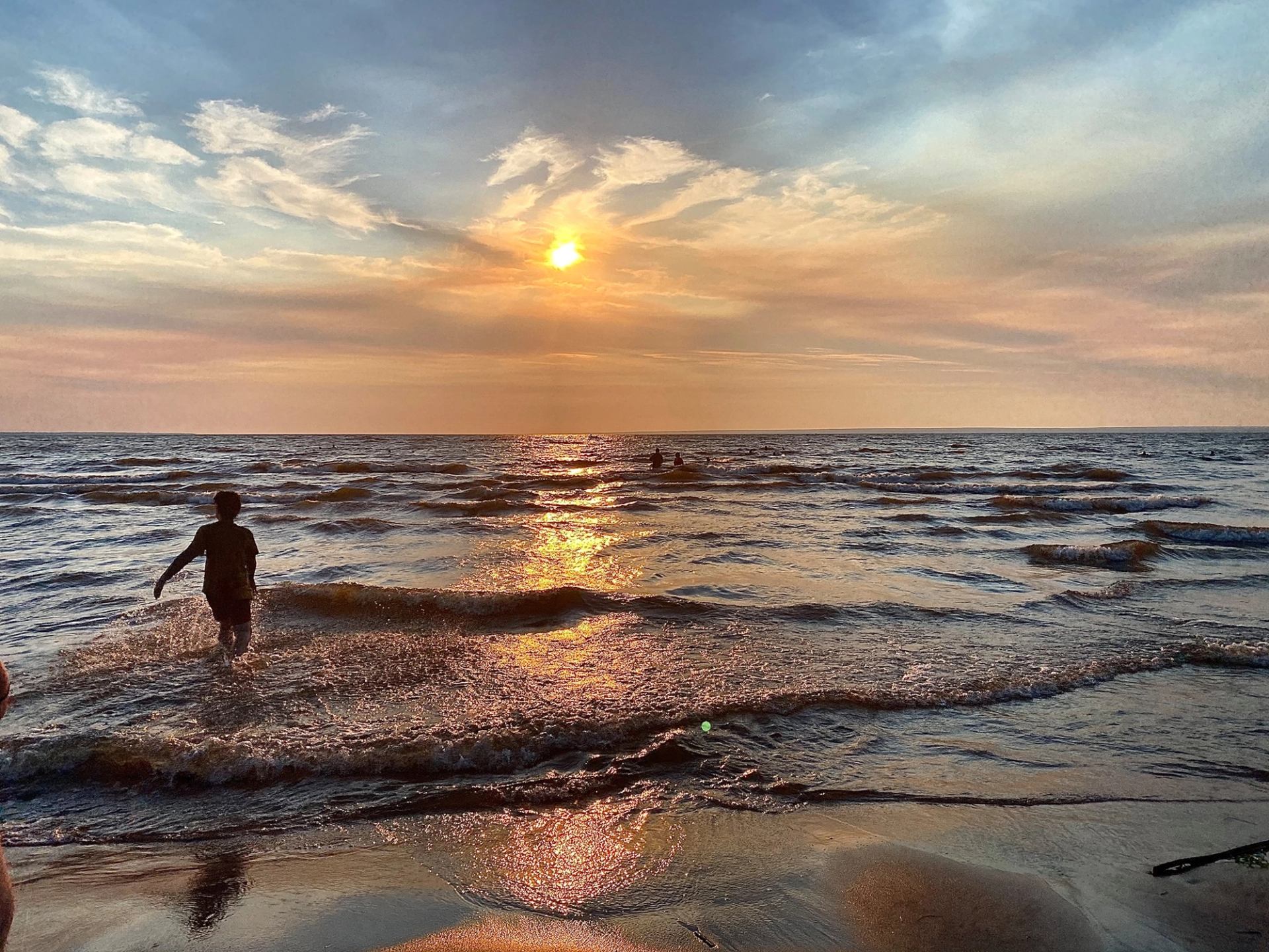 Person wades through shoreline waves beneath a cloud-streaked sunset.
