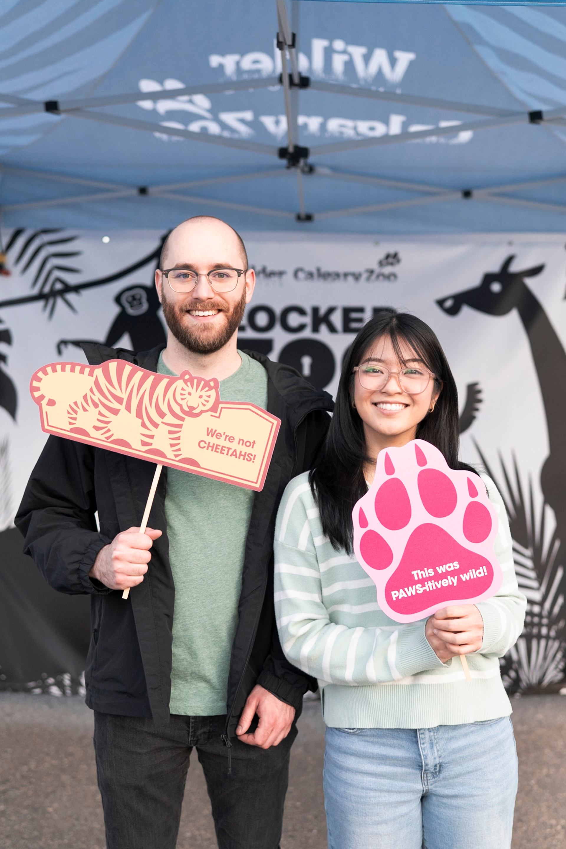 Two people hold animal pun signs under a Wilder Institute Calgary Zoo canopy.