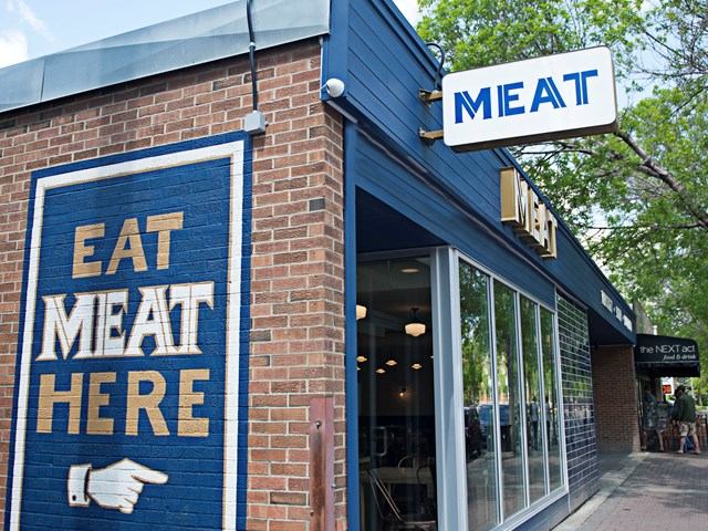 Brick restaurant exterior with “EAT MEAT HERE” sign and large windows.