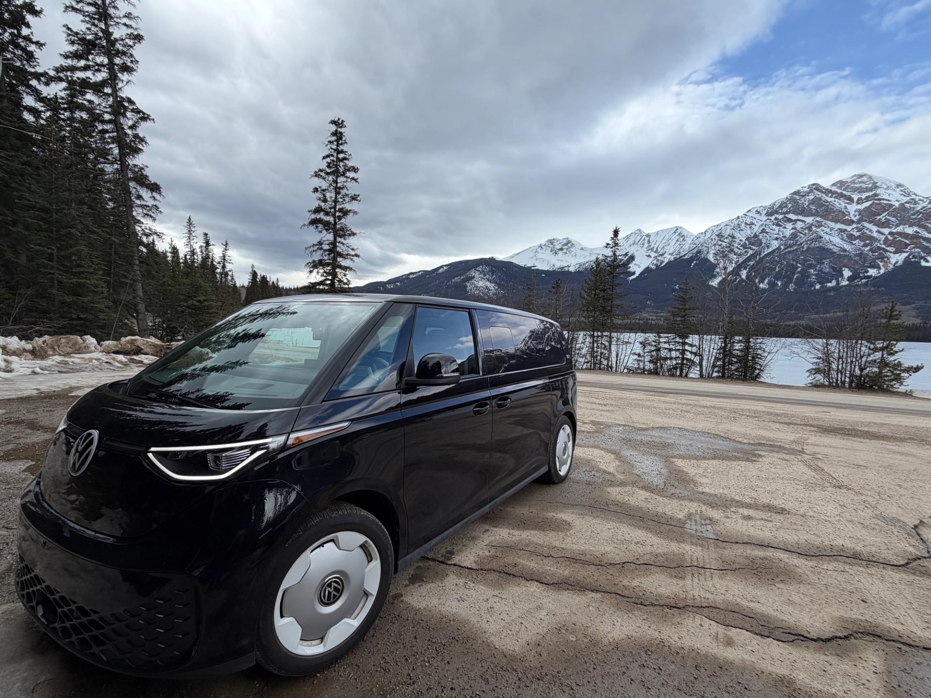 Black Volkswagen van parked by frozen lake with snow-capped mountains and evergreen trees.