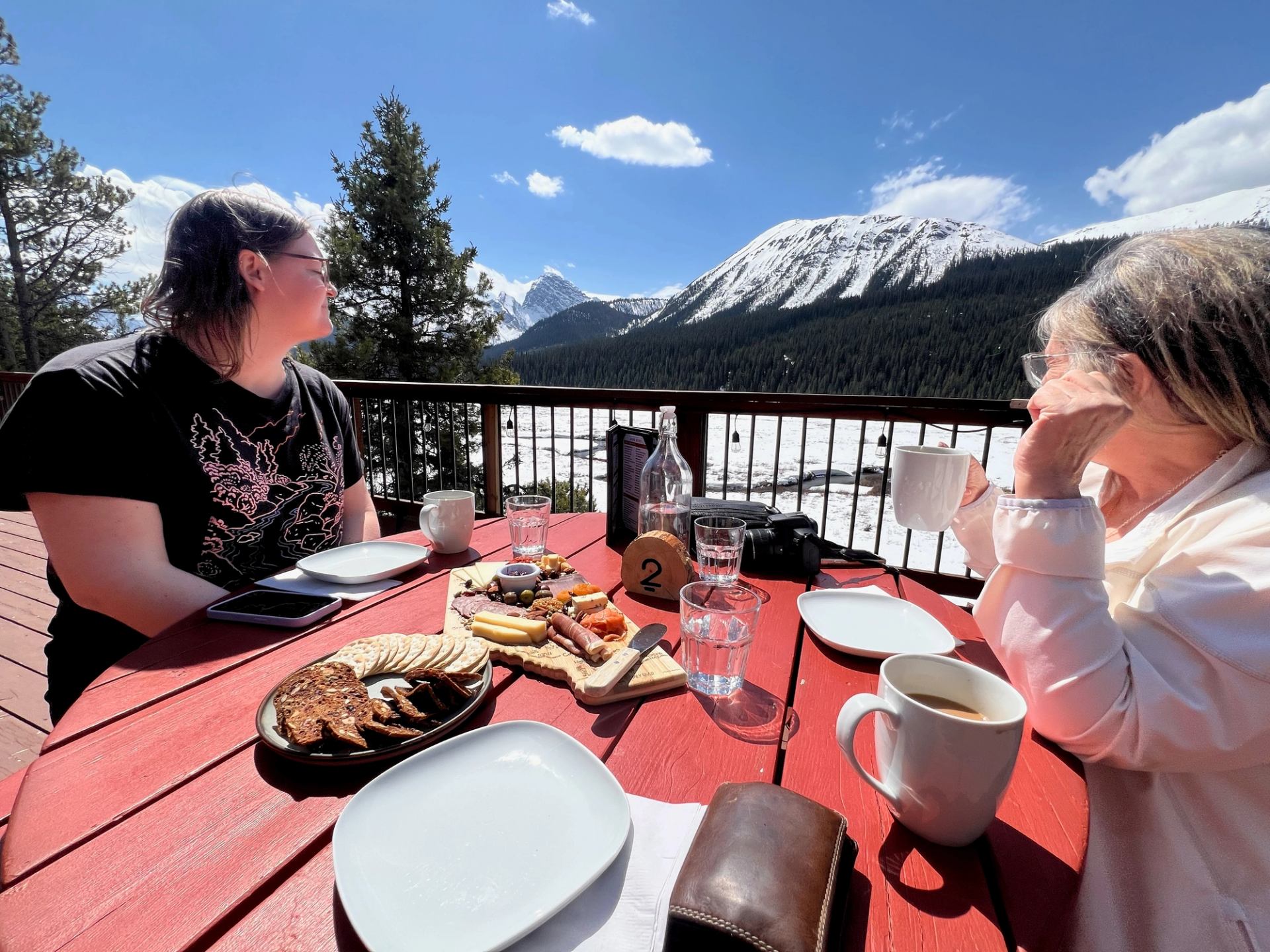 Two people seated at an outdoor table enjoying coffee and snacks with snowy mountain views.