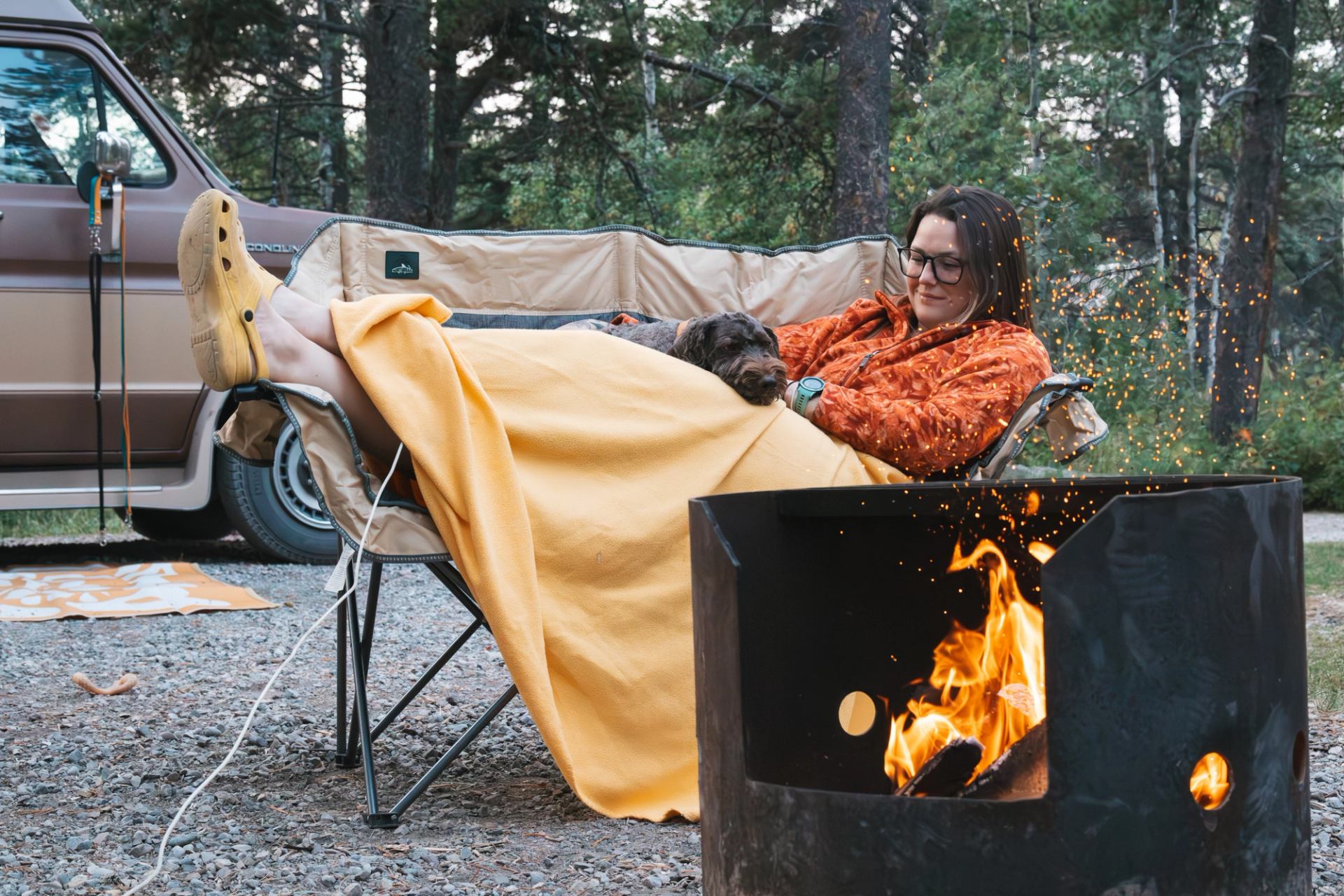 Person in camp chair under yellow blanket with dog beside fire pit and van.
