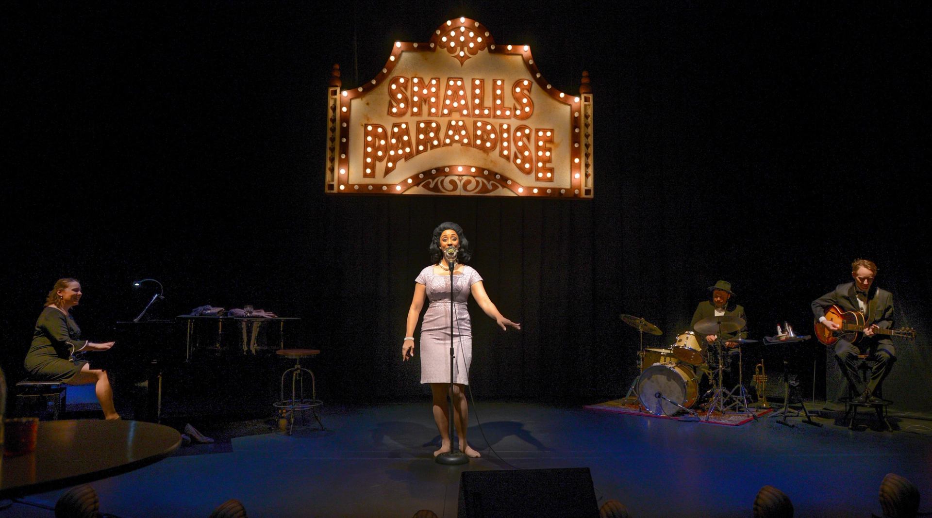Singer performs on stage with pianist, drummer and guitarist under Smalls Paradise sign.