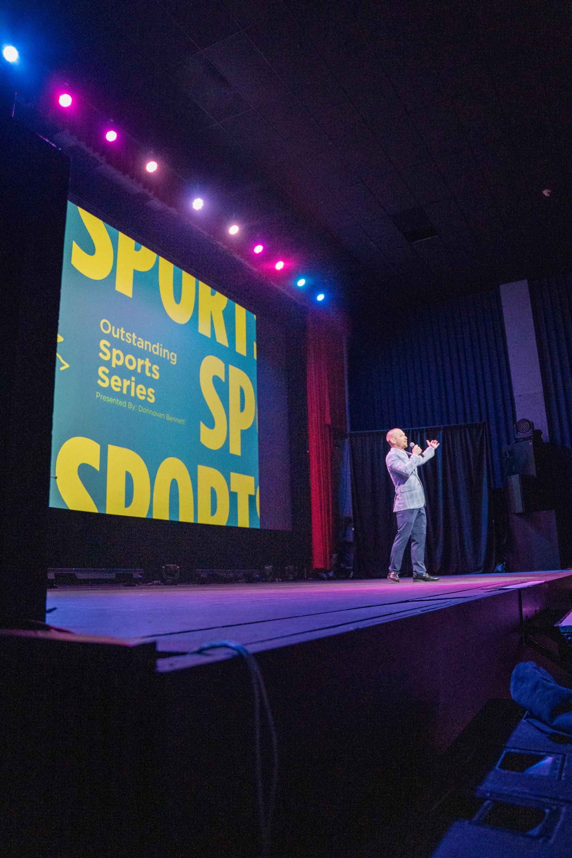 Presenter on theatre stage speaks beside large screen reading Outstanding Sports Series.