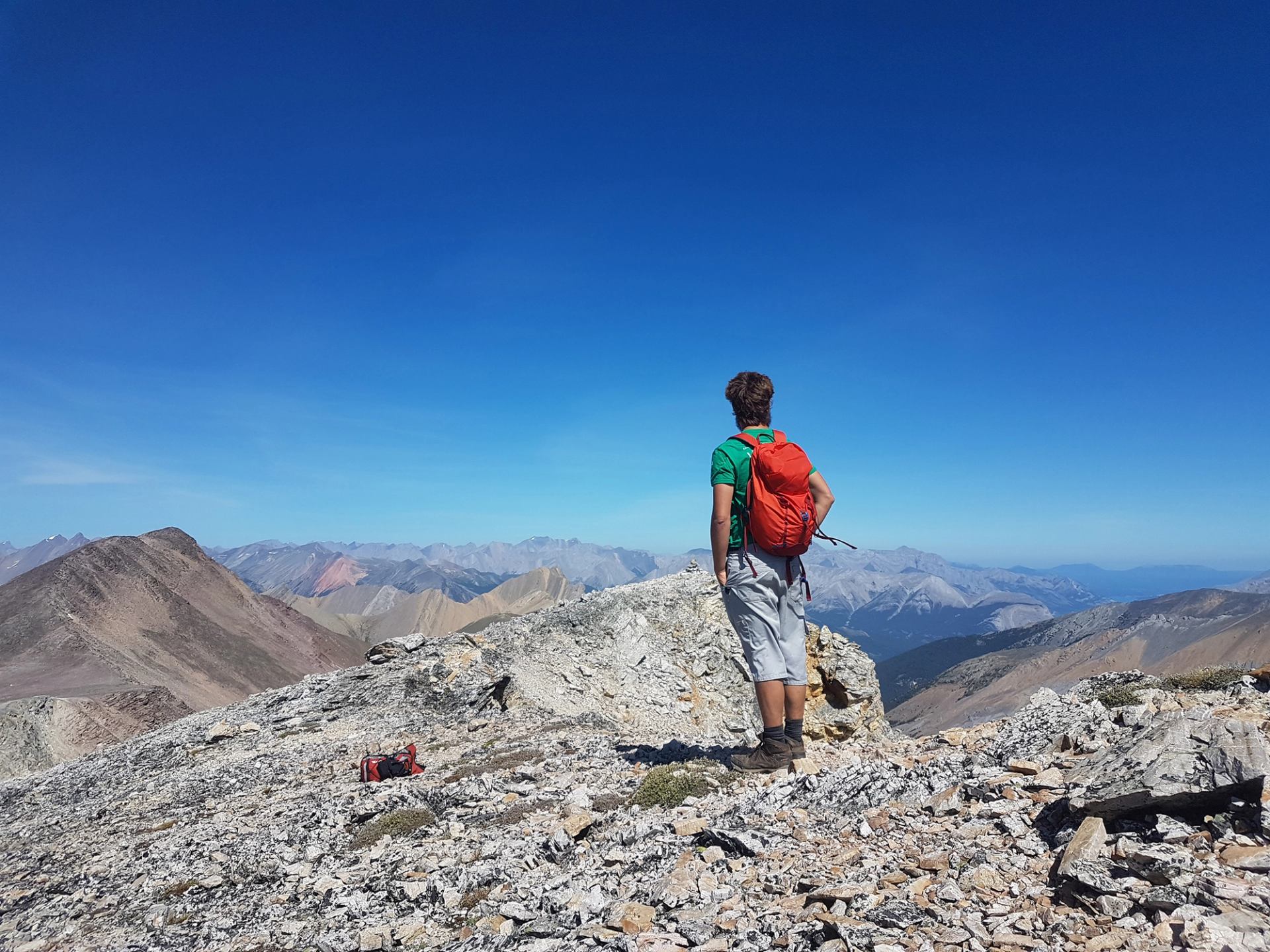 Hiker with red backpack stands on rocky mountain ridge overlooking distant peaks.