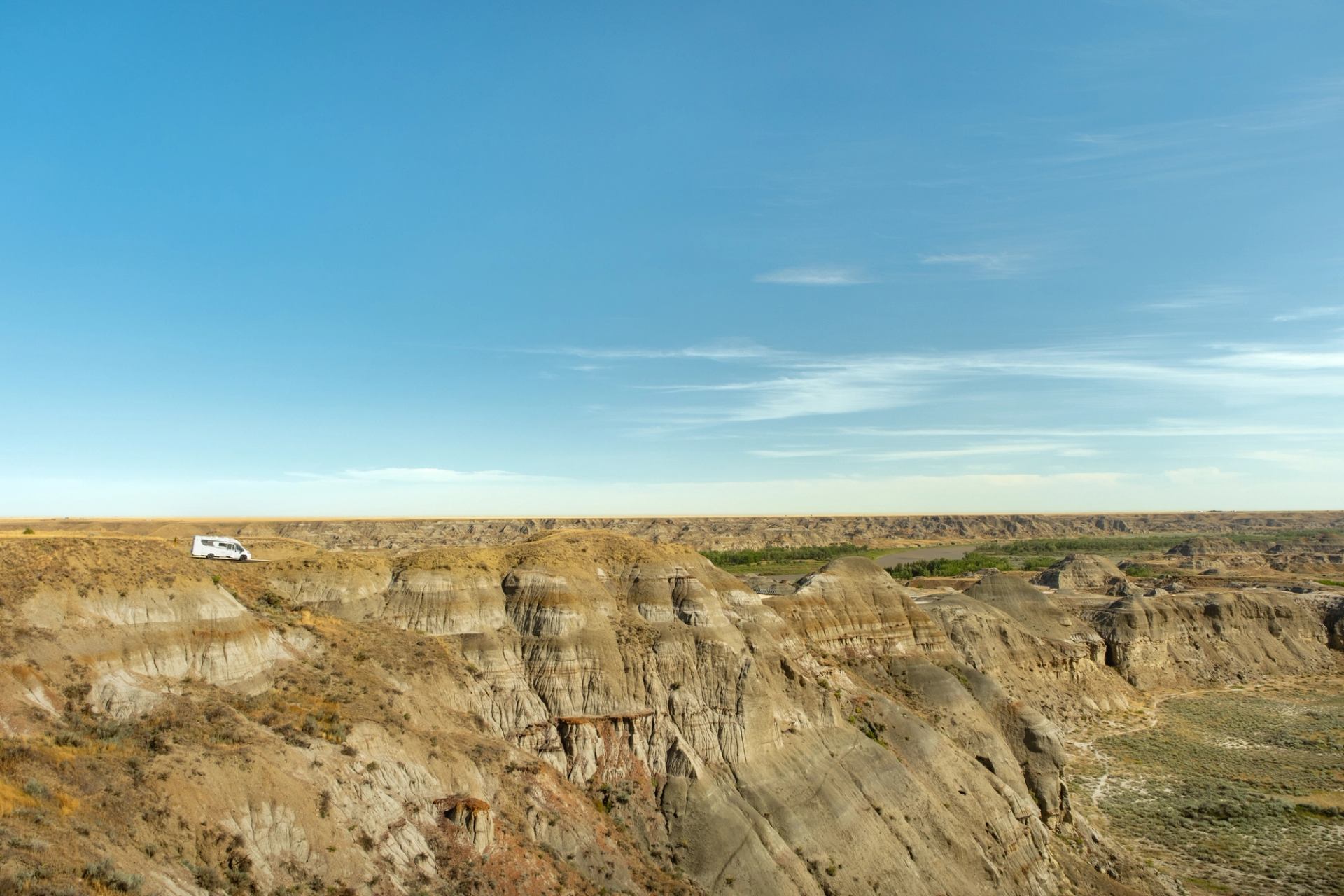 A white tour bus parked on a ridge overlooking the expansive badlands of Dinosaur Provincial Park, with layered rock formations, steep cliffs, and a wide blue sky stretching across the horizon.