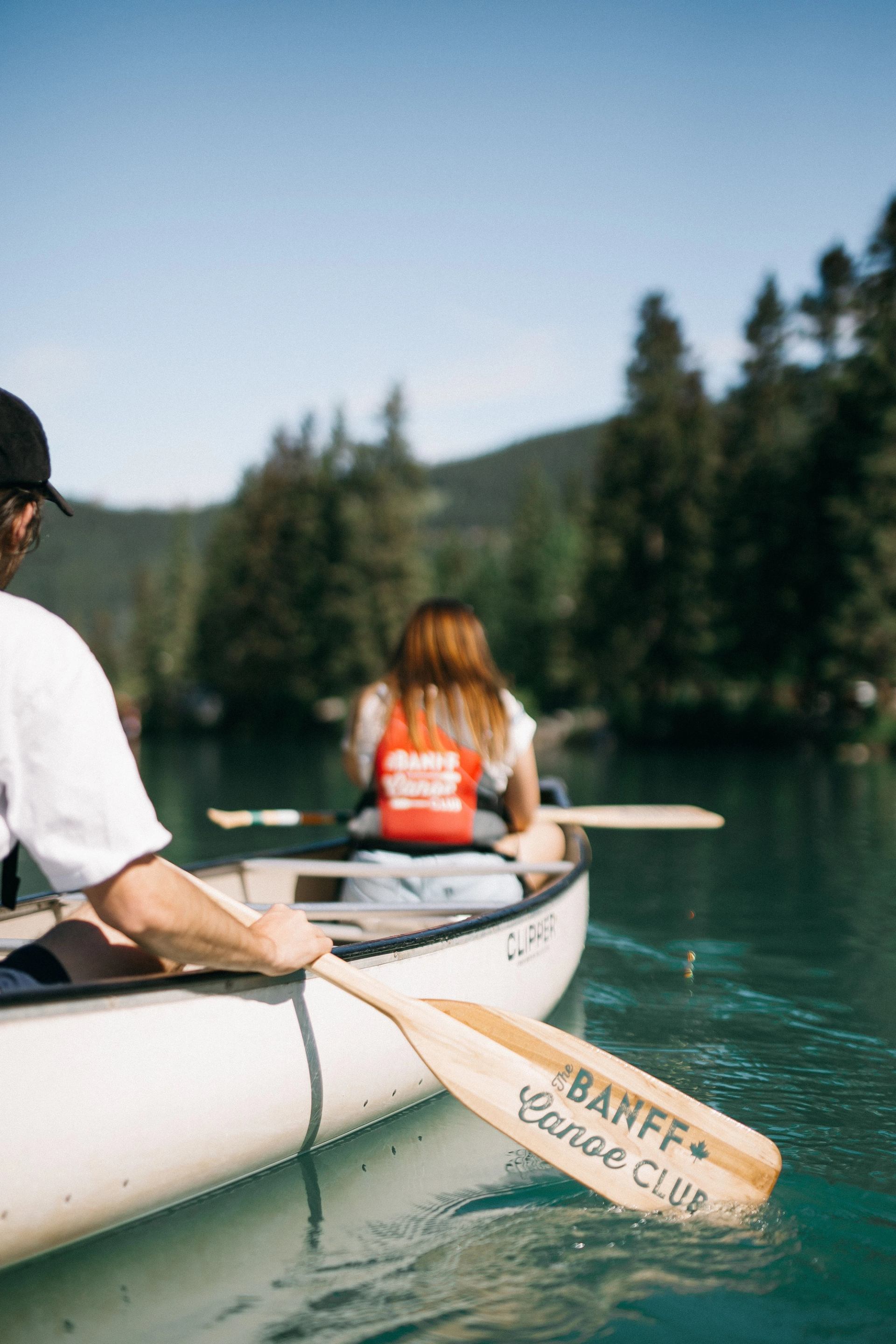 A couple paddles a canoe from the Banff Canoe Club on a calm blue lake with forested hills ahead.