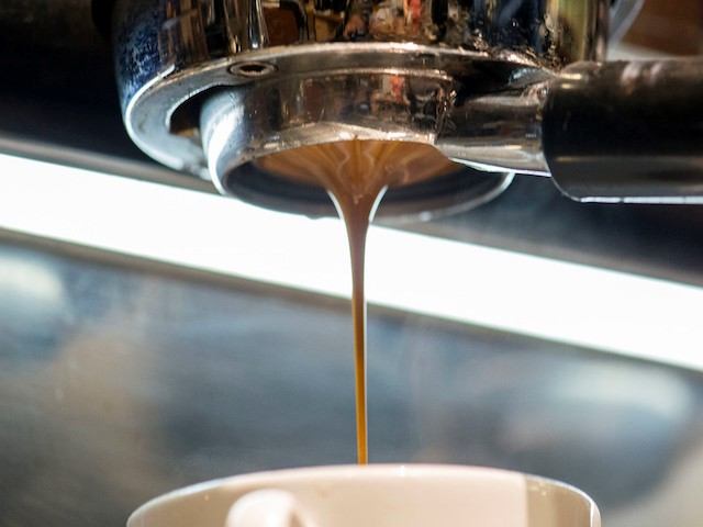 Espresso flows from a coffee machine portafilter into a cup below in a thin, steady stream. The surrounding metal surfaces reflect light from the café environment.