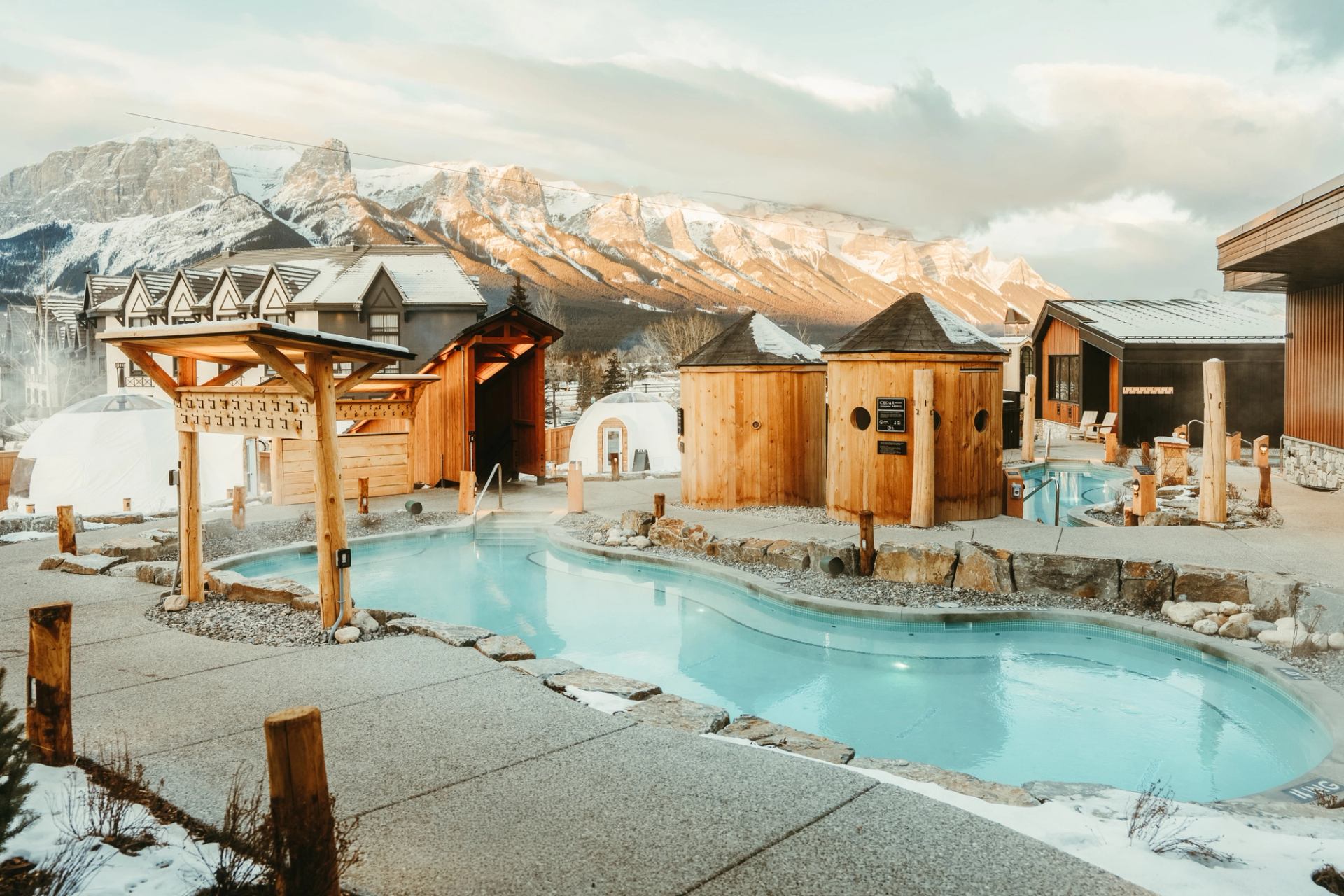 Outdoor hot pool with wooden saunas and domes, backed by snow-covered mountain range