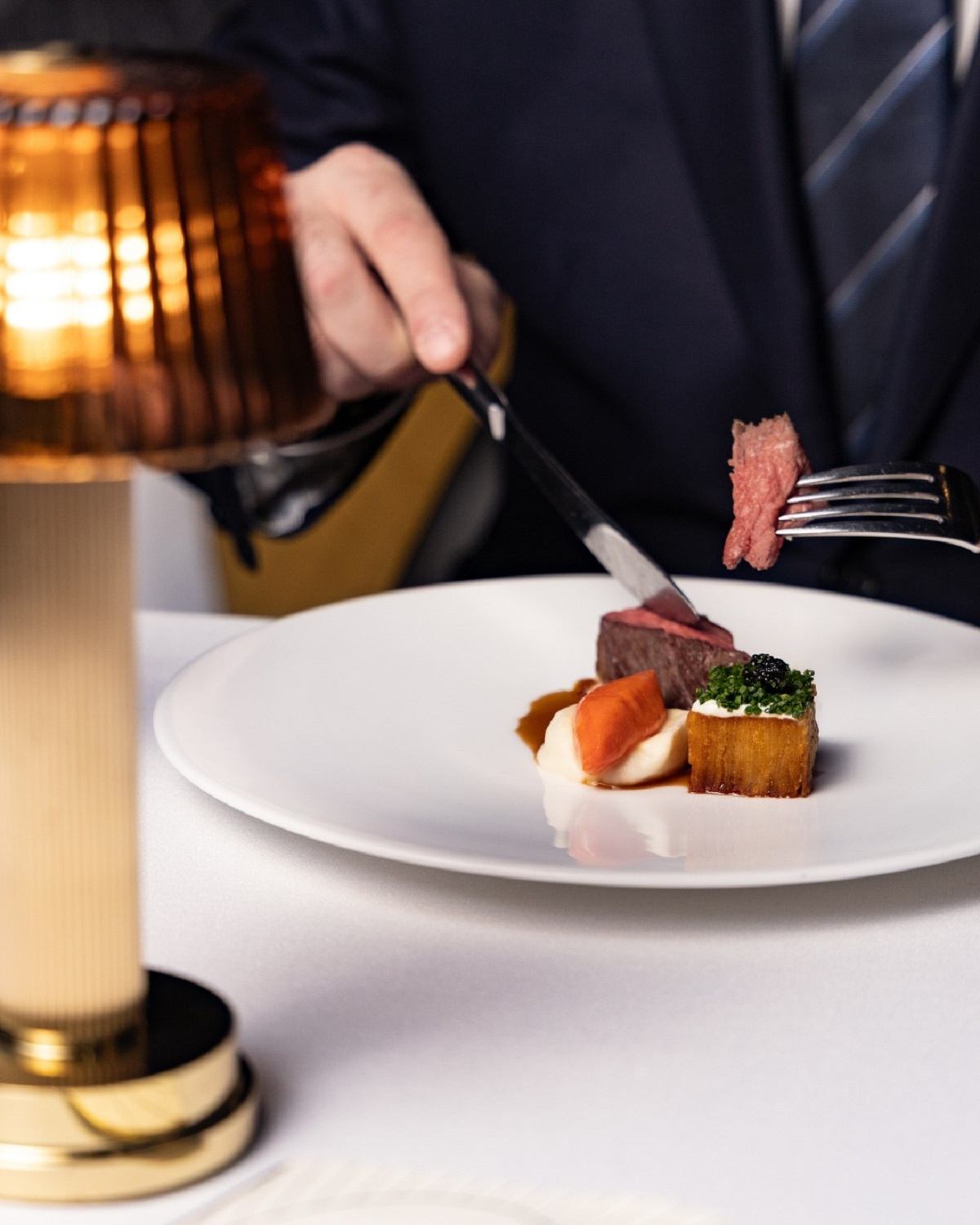 Person in suit cuts plated steak with vegetables beside a table lamp.