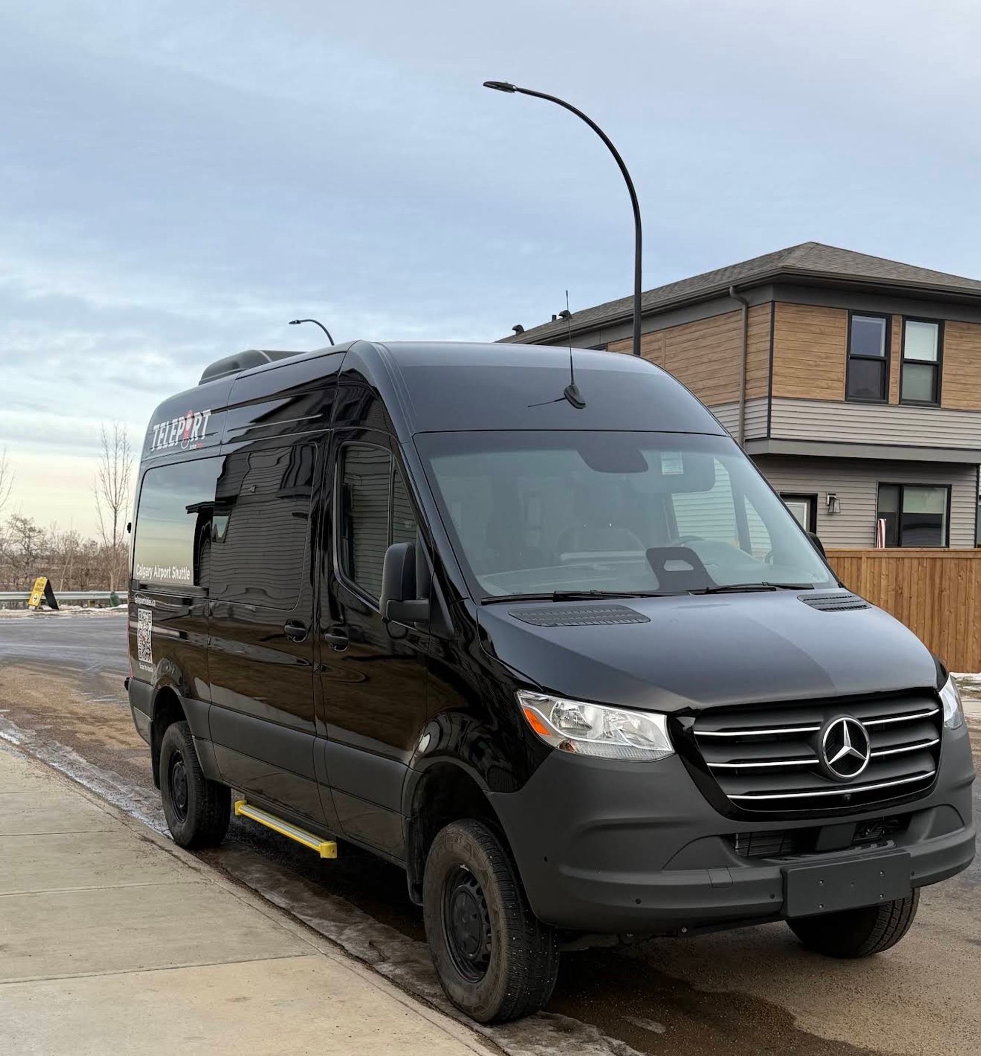 Black Mercedes-Benz shuttle van parked on residential street beside modern two-storey houses.
