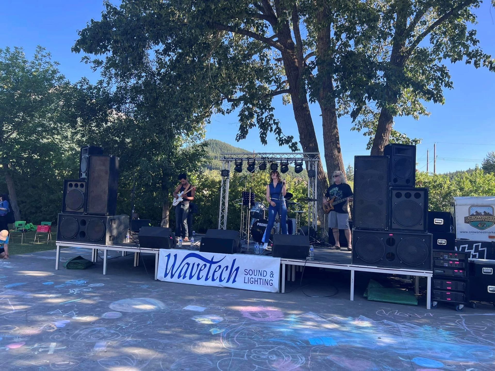 Outdoor band performing on stage with large speakers, trees and chalk drawings visible.
