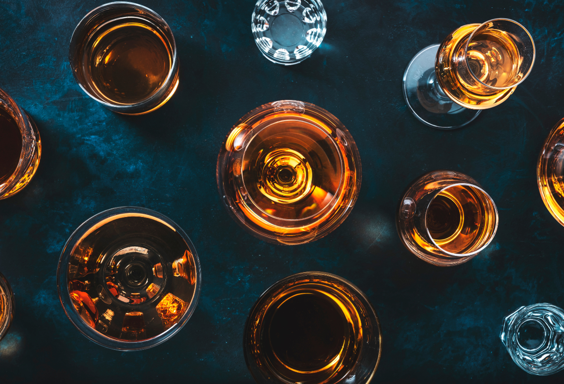 Assorted glasses of amber liquor arranged on a dark blue surface.