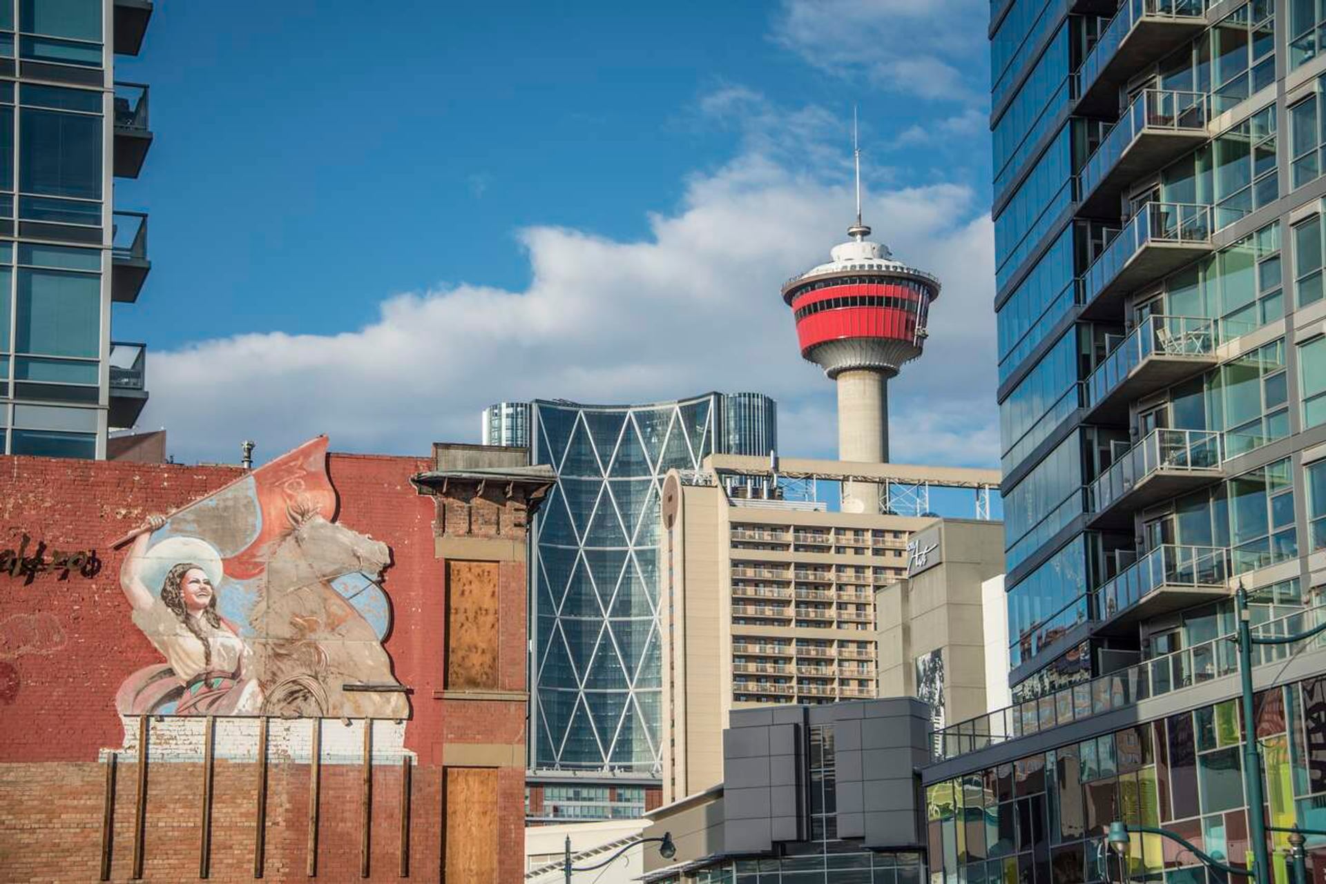 Calgary Tower rises above downtown buildings and mural on brick wall under blue sky.