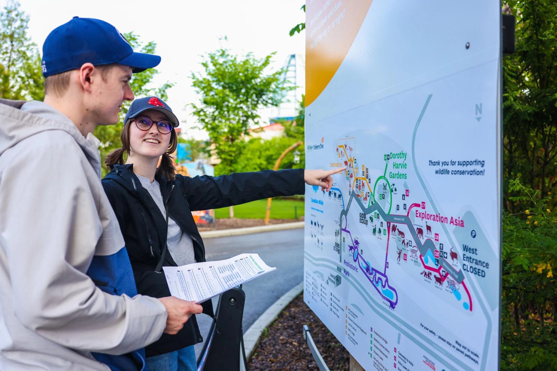 Visitors in caps study an outdoor zoo map, one person pointing at destinations.