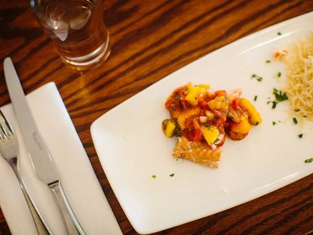 Plated salmon topped with mango salsa and rice on a white dish, beside cutlery and a glass of water.