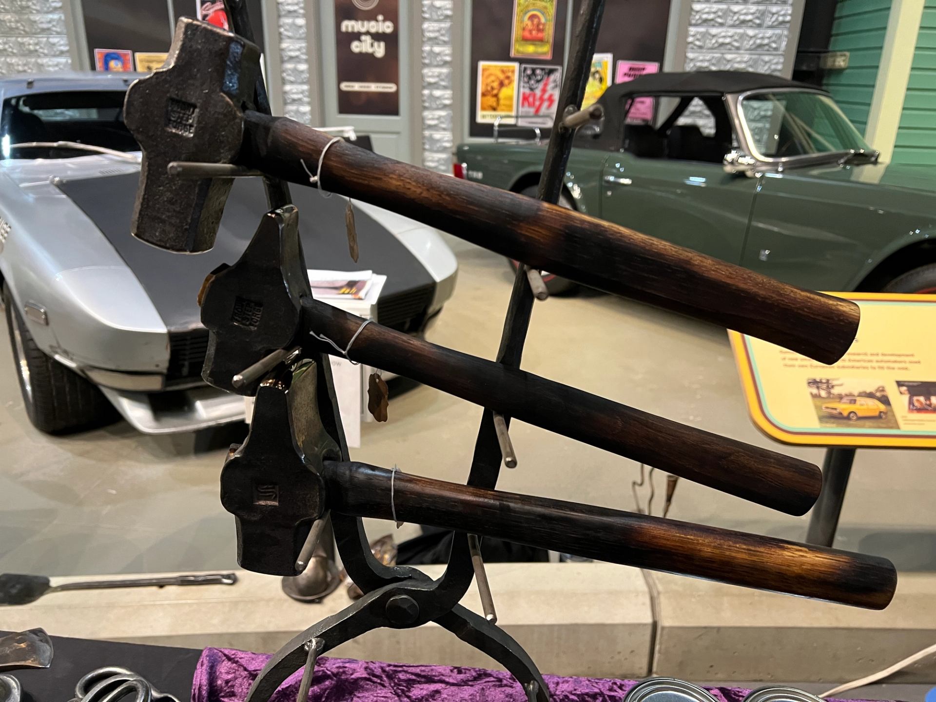 Metal display stand holding wooden-handled hammers, with classic cars in background.