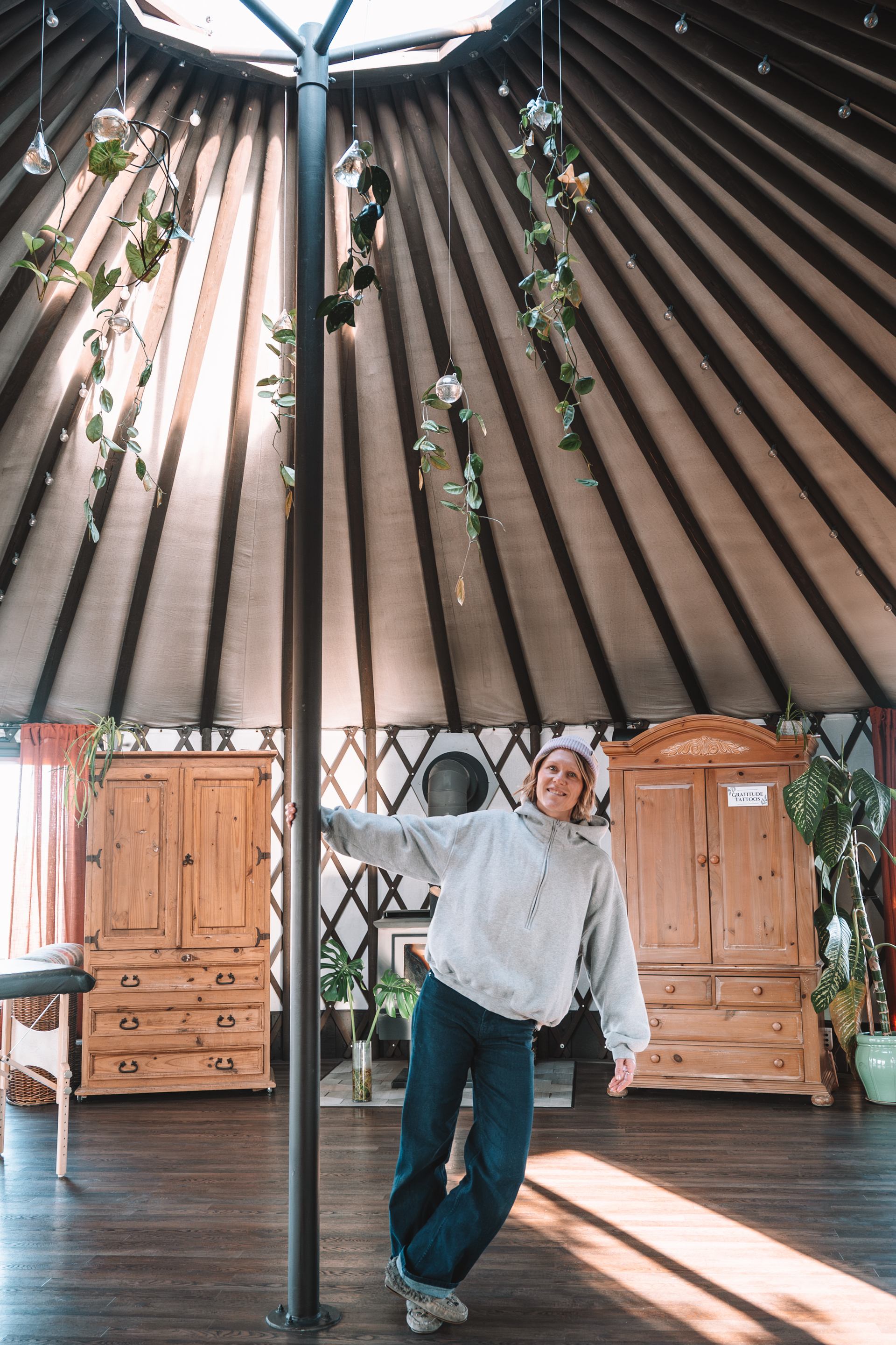 Person holds a central support pole inside a yurt with hanging plants and wooden cabinets.