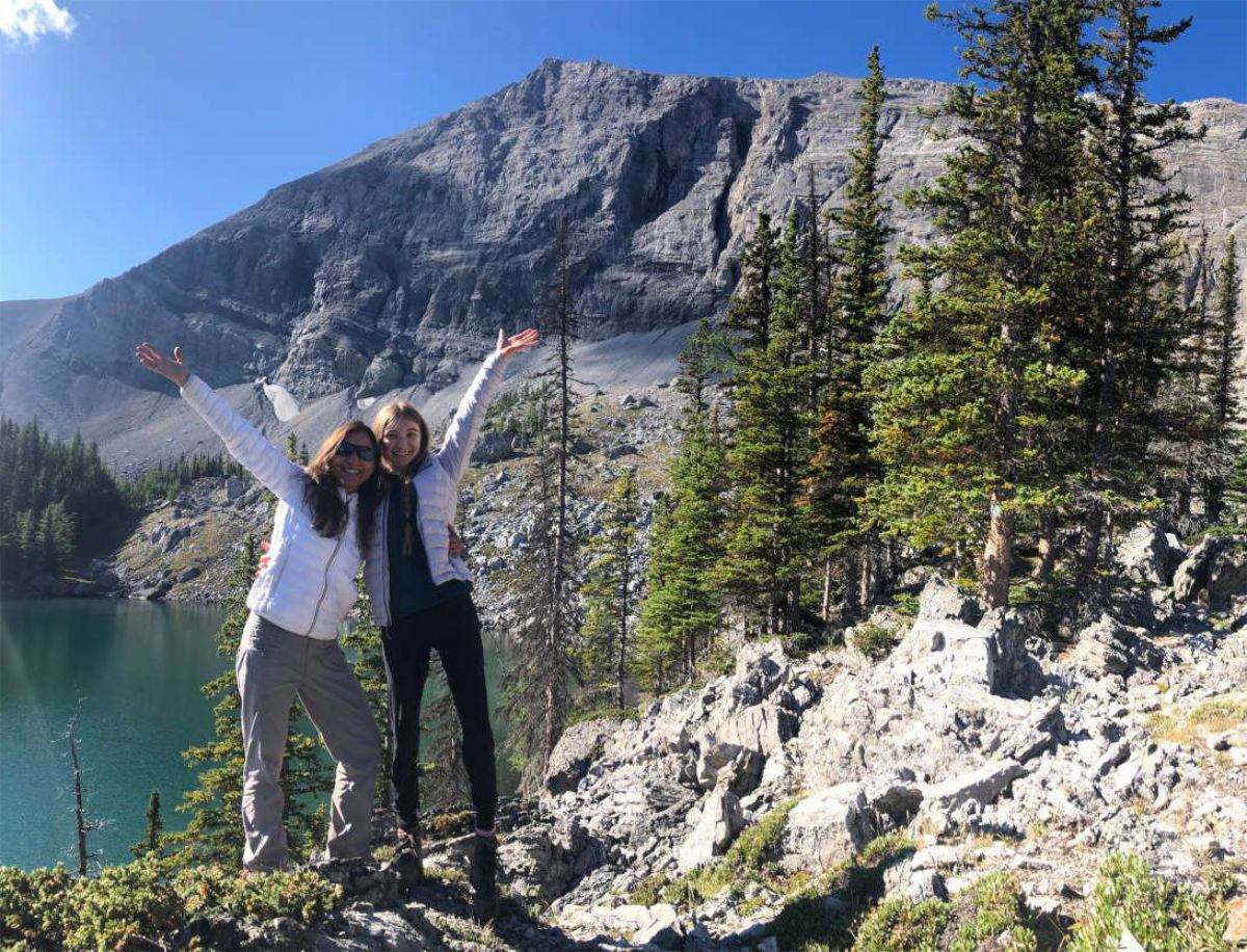 Two hikers stand on rocky lakeshore with evergreen trees and steep grey mountain behind.