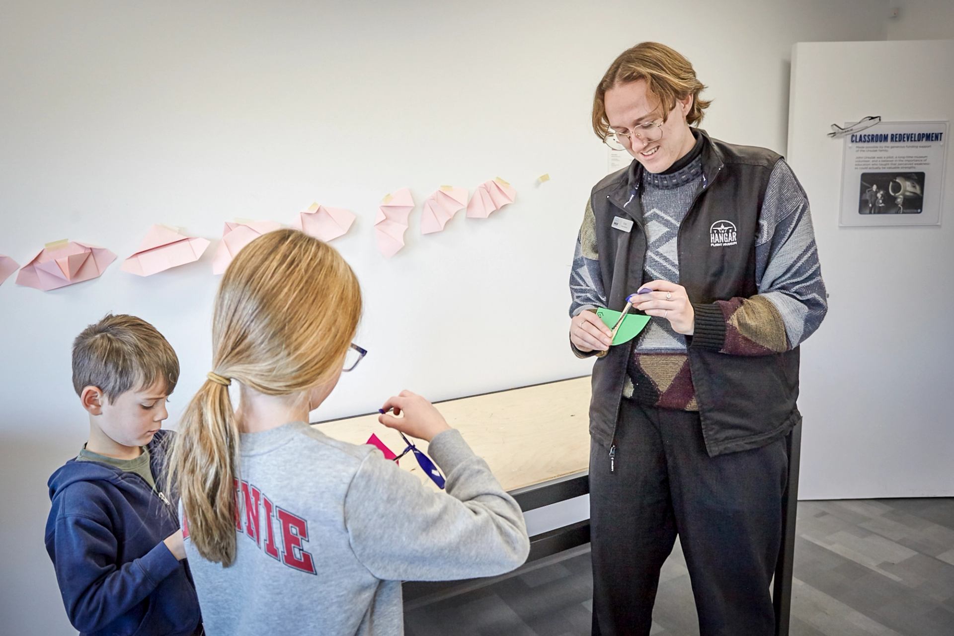 Museum educator helps children make paper aircraft at classroom table on display wall.