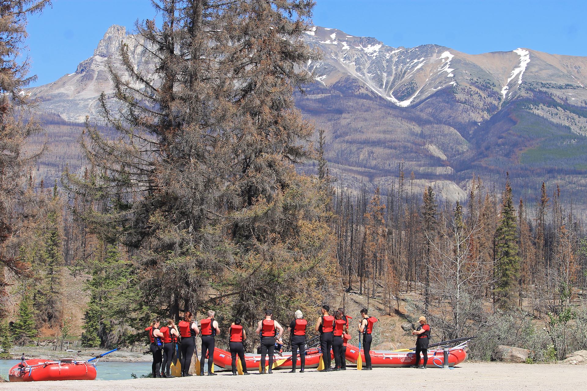 A group in red life vests stands by a river with rafts, surrounded by fire-damaged trees and snowy mountains.