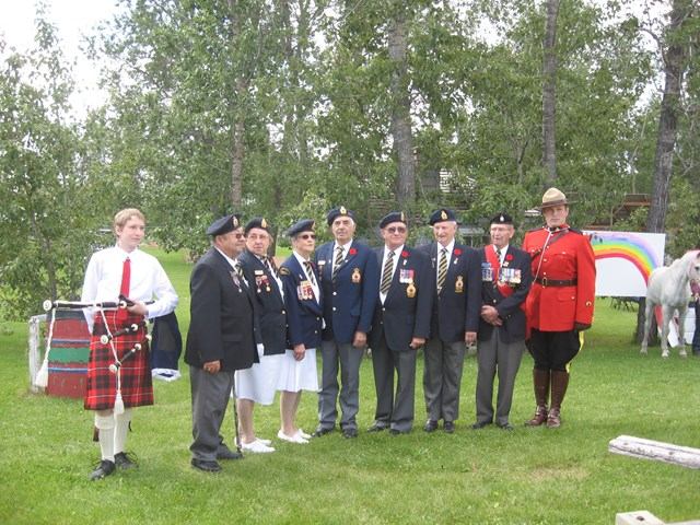 Group of uniformed veterans and a Mountie posing outdoors beside a bagpiper and rainbow mural.