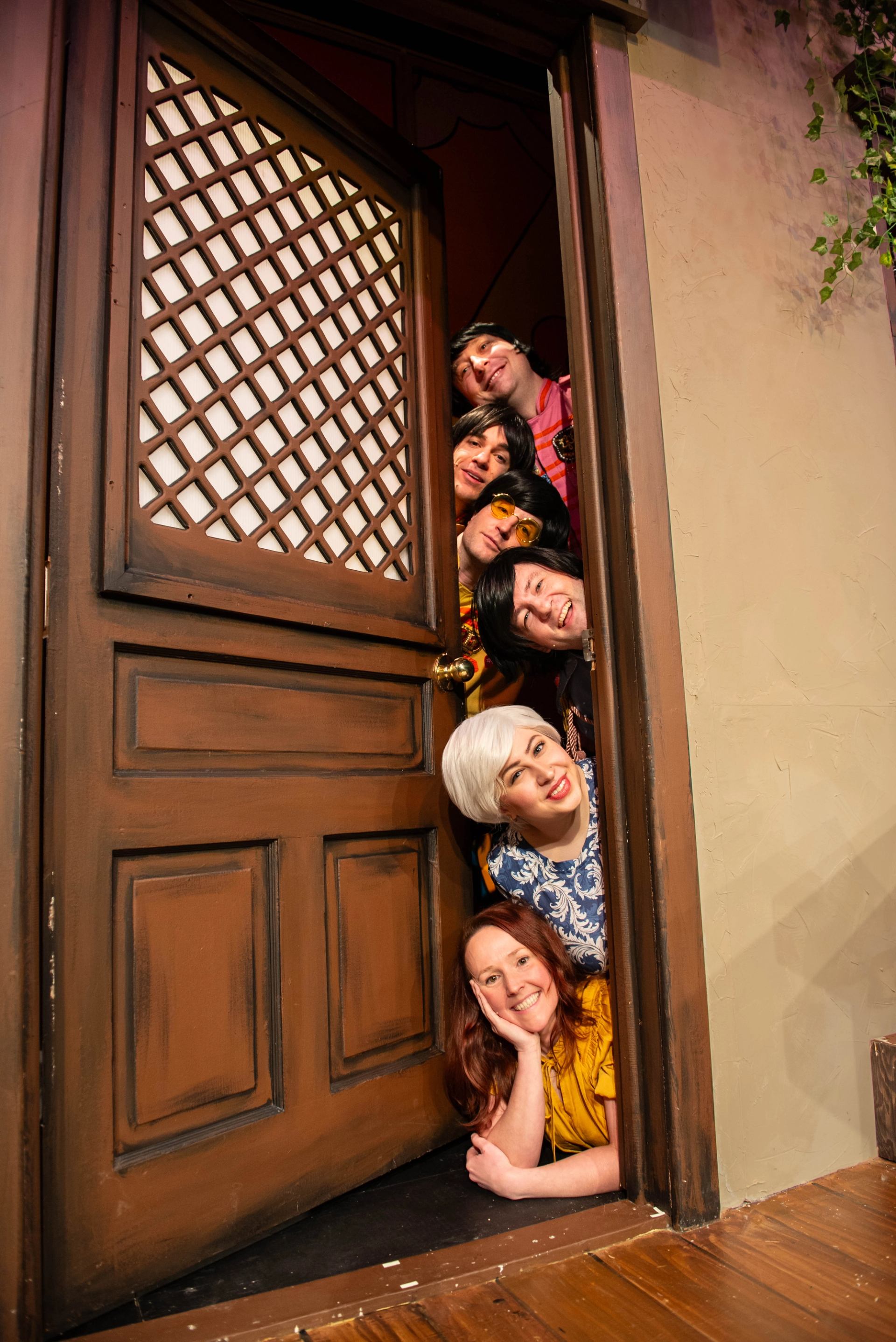 Six people in colourful costumes lean through a wooden stage door, stacked vertically.