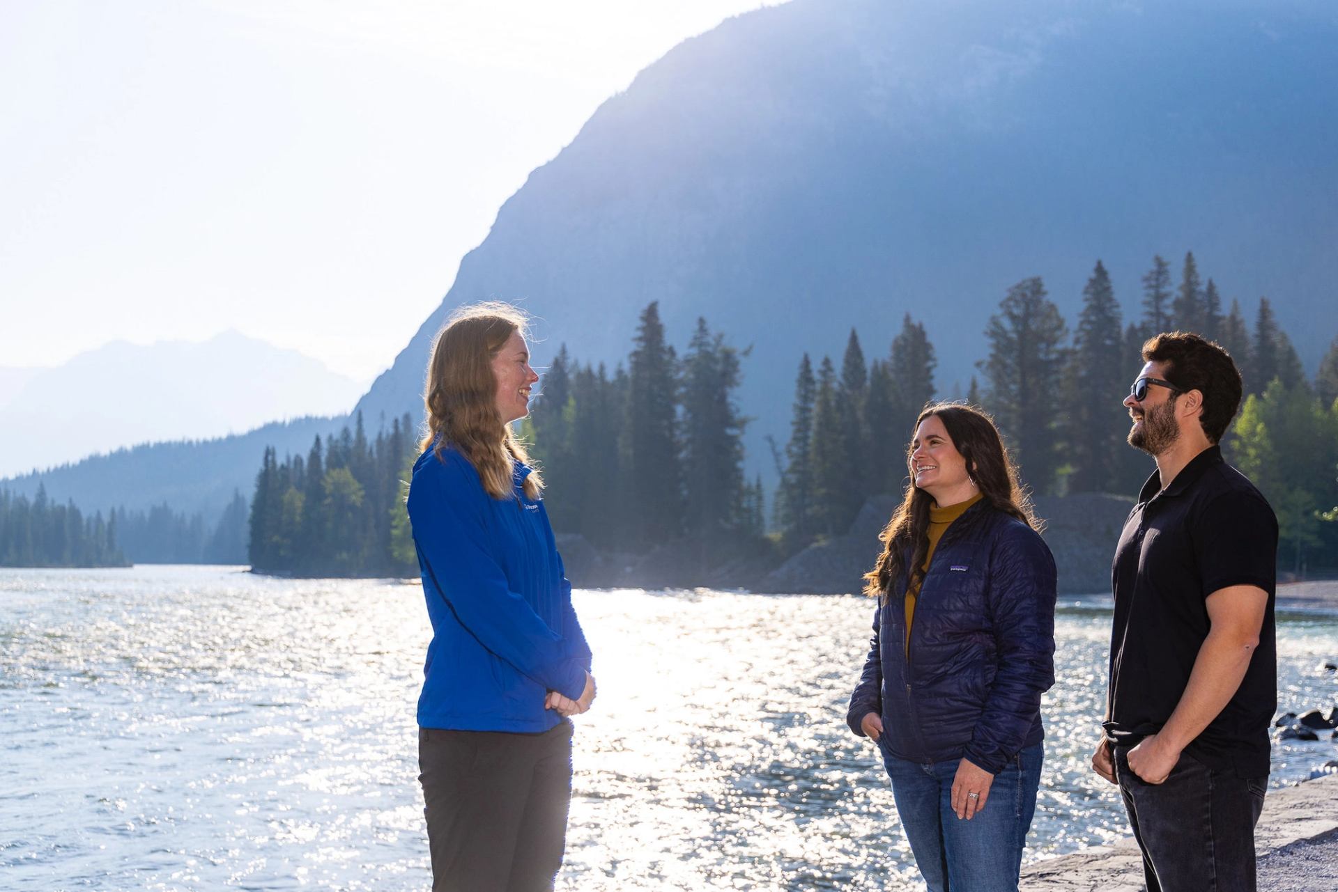 Three people stand talking beside a shimmering lake with forested mountains behind.