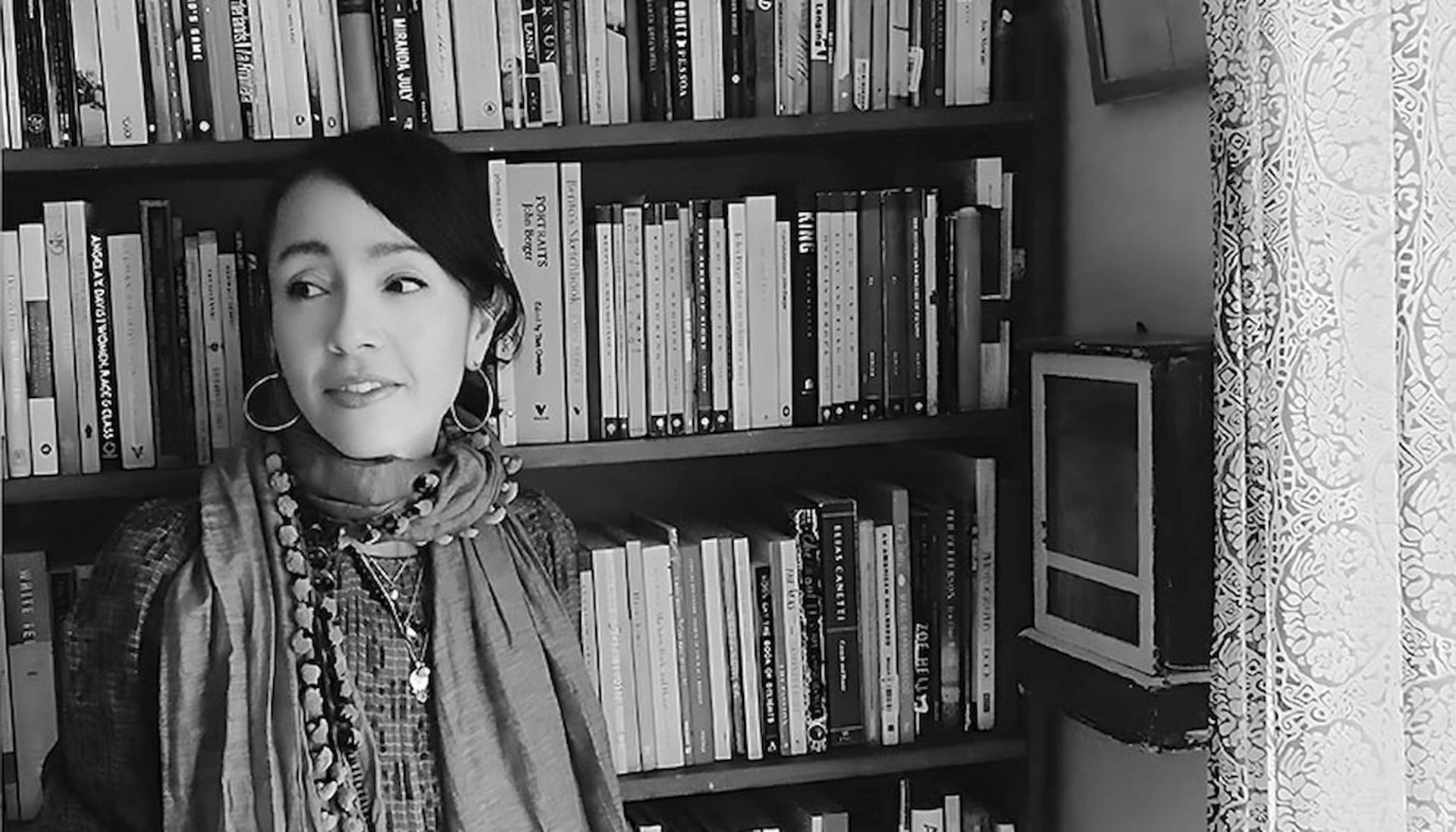 Person wearing scarf and jewellery stands by full bookshelf beside patterned curtain.