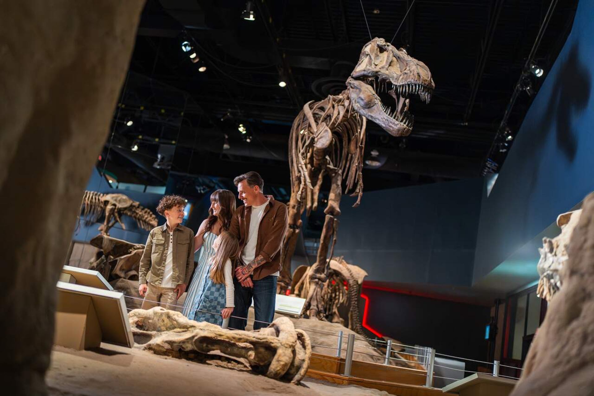 Visitors stand near a large Tyrannosaurus rex skeleton in a dim museum gallery.