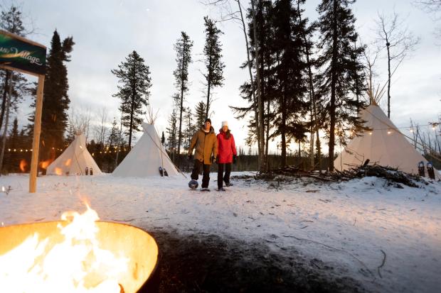 A couple snowshoeing towards a bon fire, tipis in the background, at Aurora Borealis Indigenous Village.