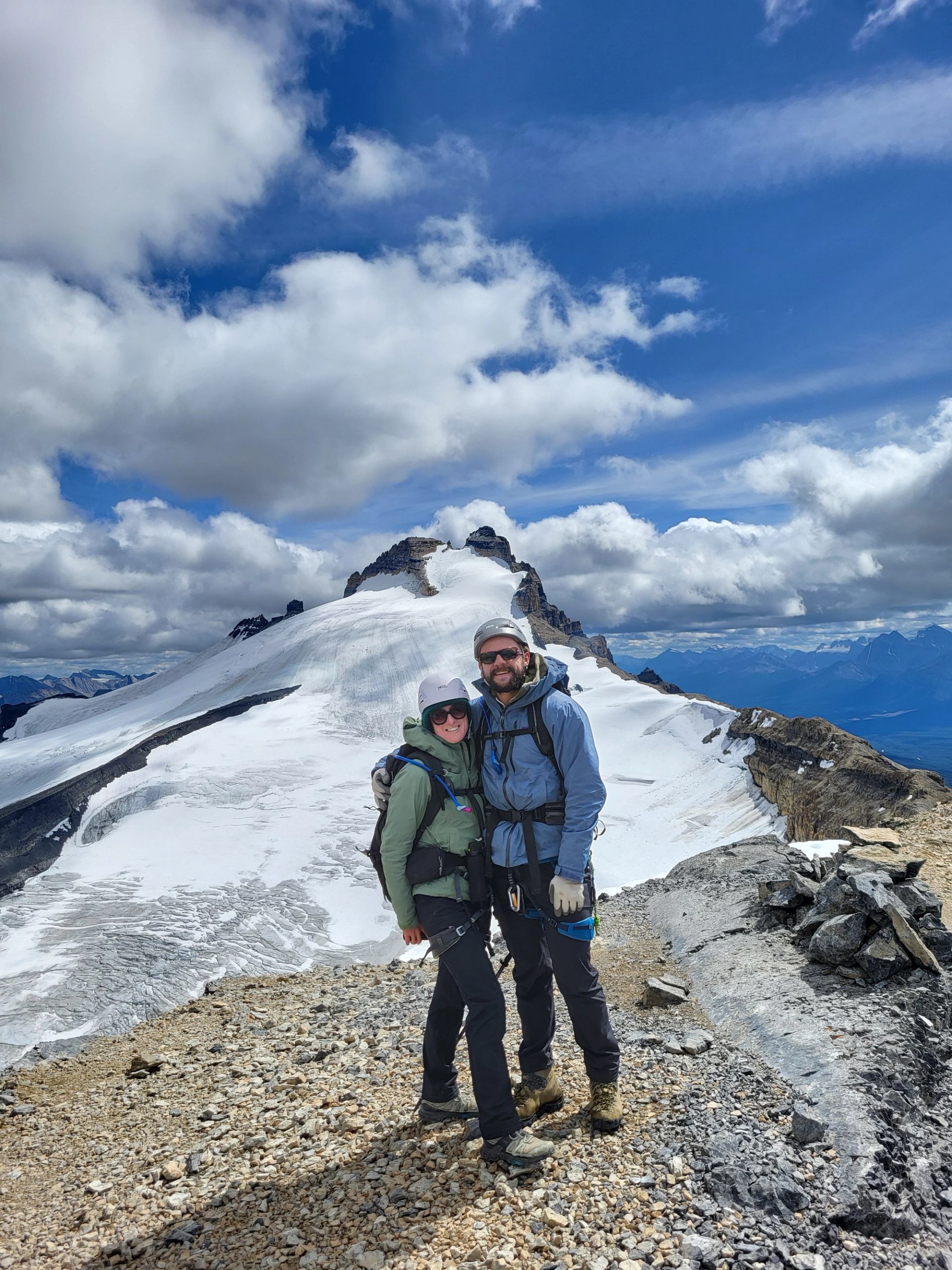 Two hikers stand on snowy mountain peak under clear sky with clouds.