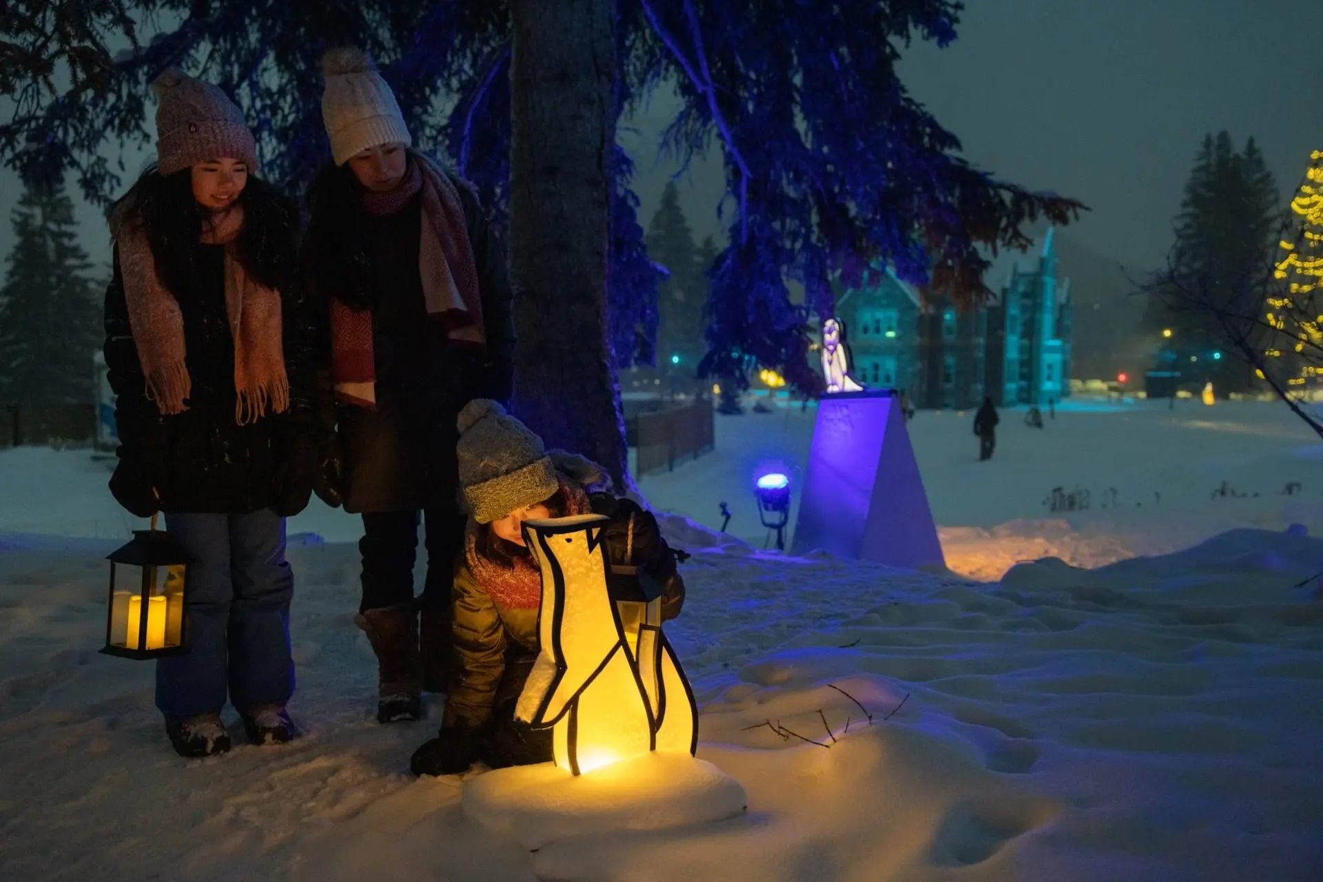 Three people explore glowing snow art with lanterns in a snowy, festive nighttime forest.