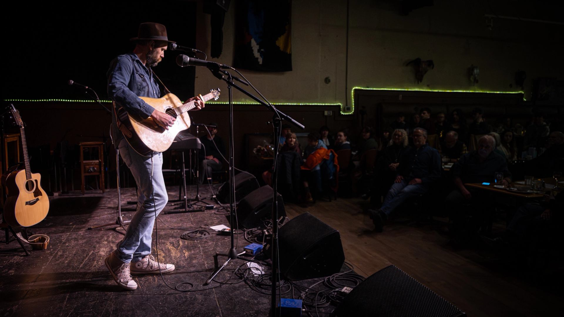 Guitarist in hat performs on small stage while seated audience watches in dim venue.