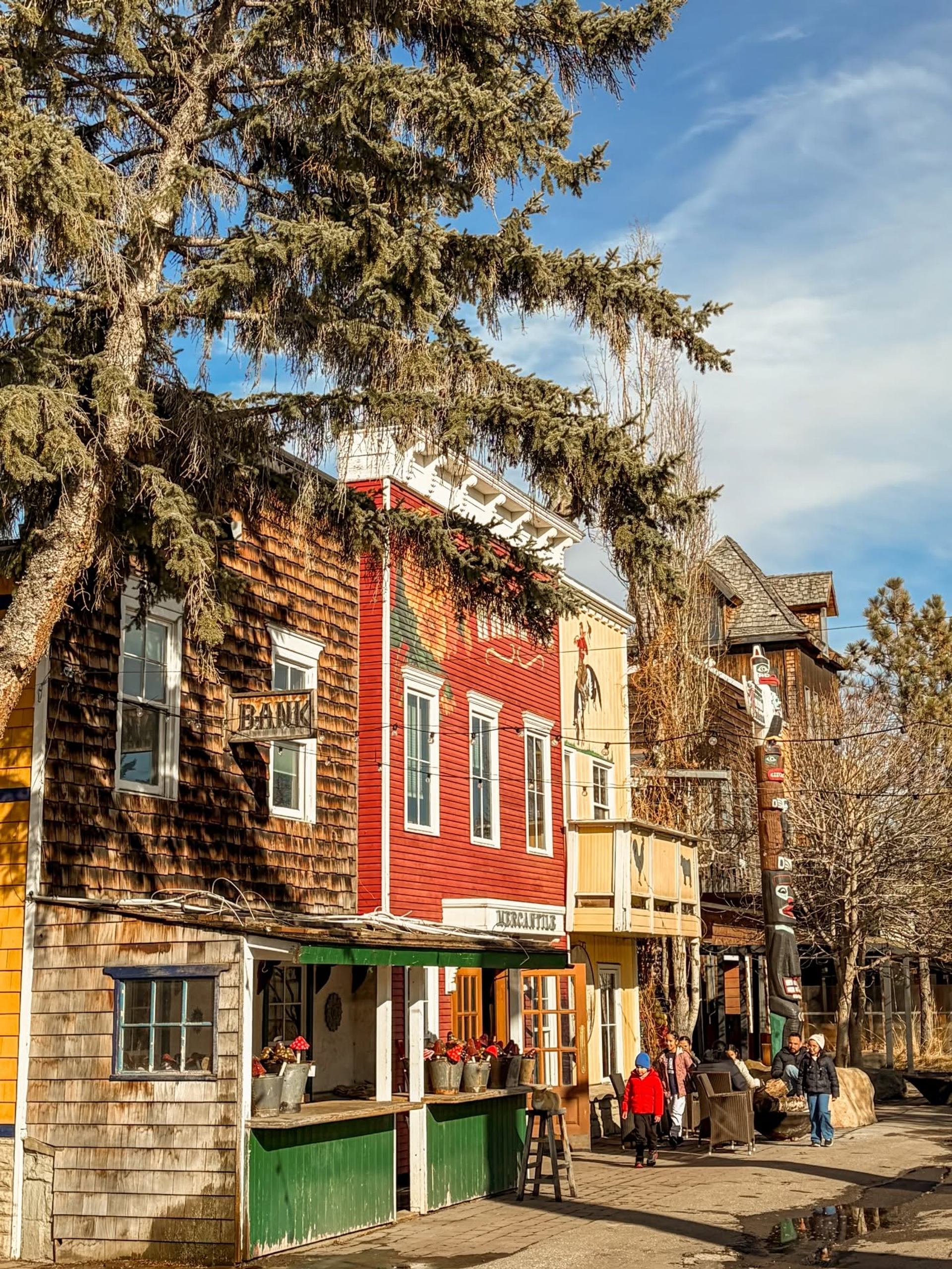 Historic-style wooden storefronts and bank sign beside tall evergreen tree and pedestrians.
