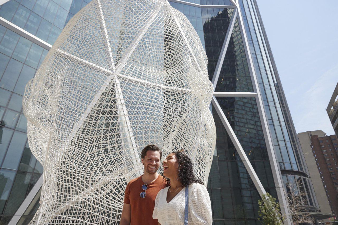 Large wire-mesh head sculpture outside glass office towers with two people standing below.