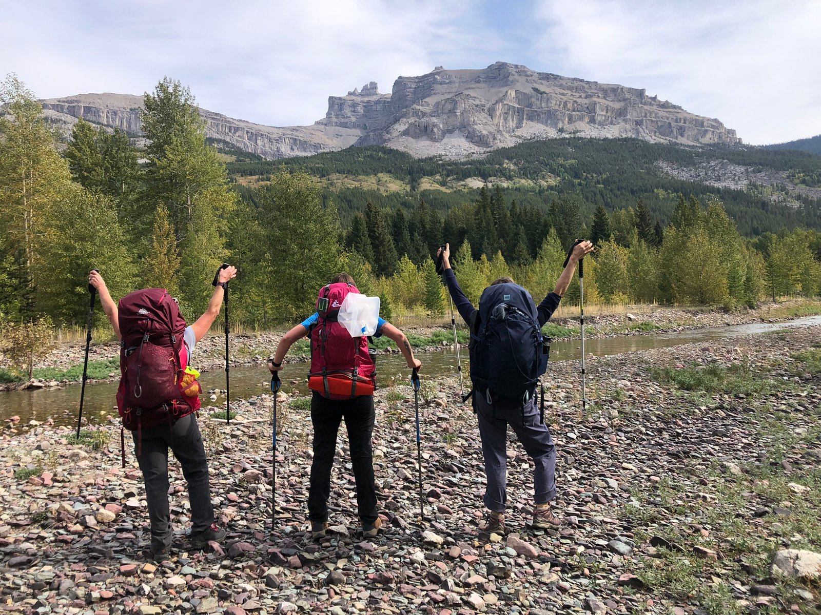 Three backpackers with trekking poles stand on rocky riverbank facing forested mountain cliffs.
