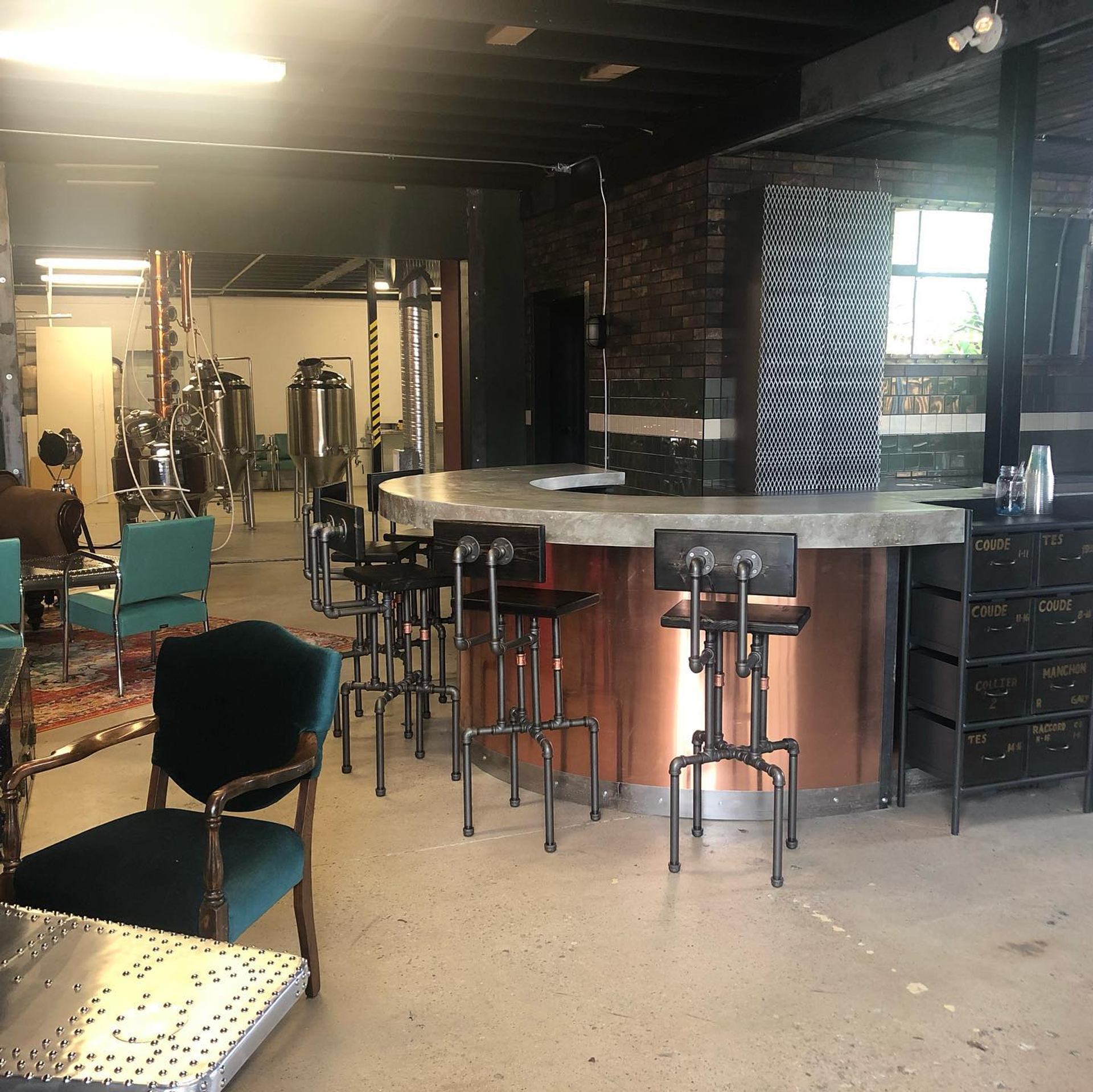 Industrial-style bar with metal stools, teal chairs and visible stainless steel brewing tanks.