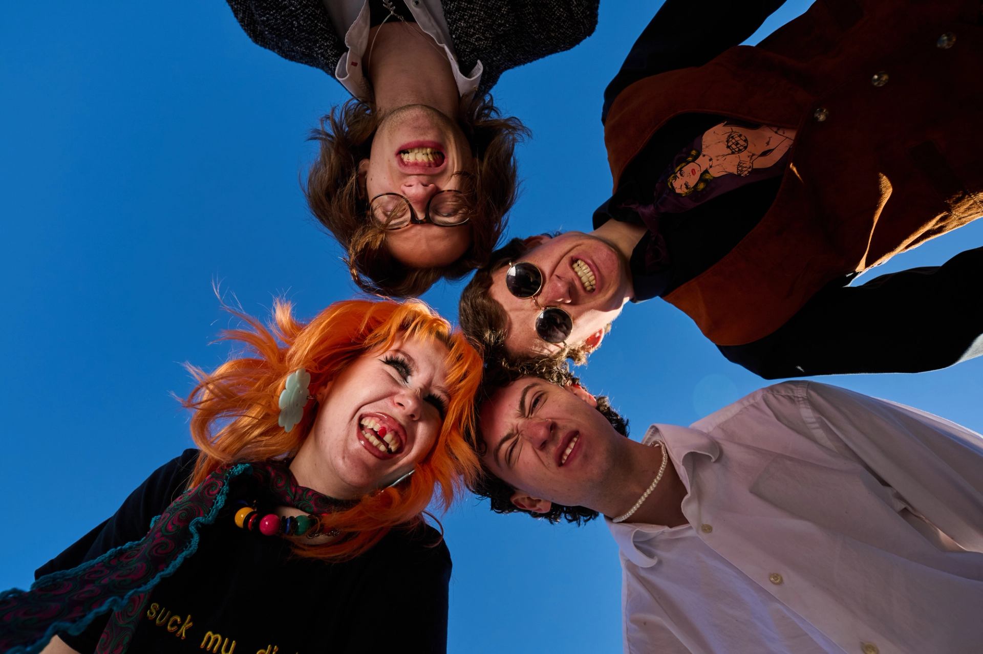 Group of four people lean over camera, making faces against clear blue sky.
