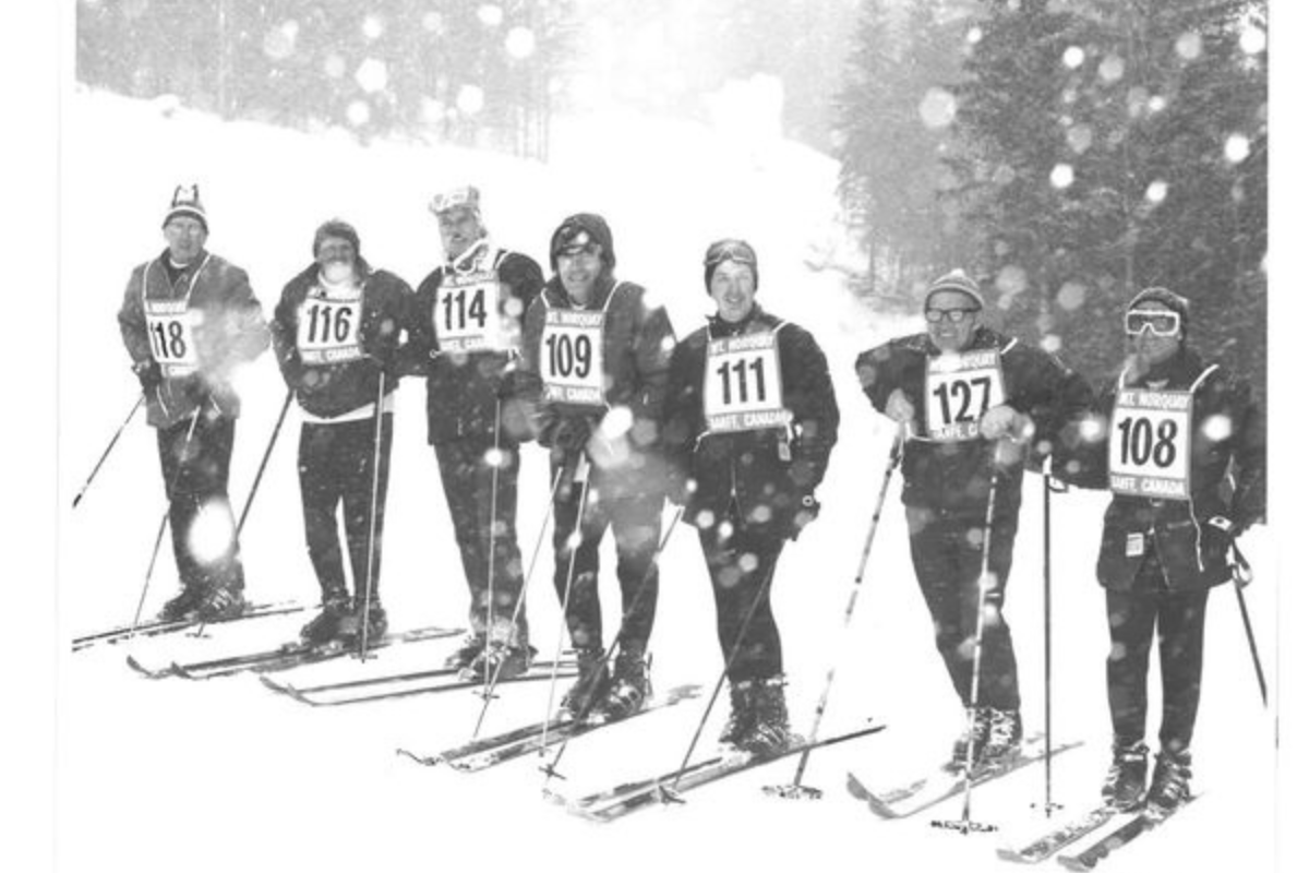 Group of skiers with numbered bibs standing on snowy trail during ski race.